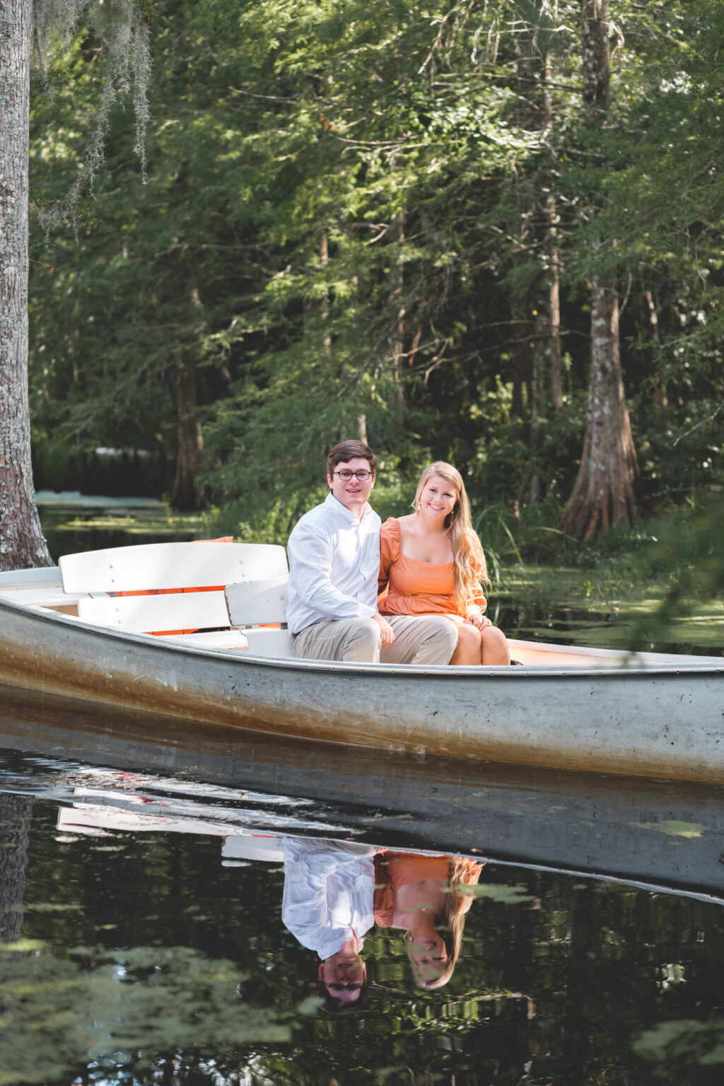 Elopement Wedding A young man in a white shirt and khaki pants and a young woman in an orange dress sit together in a small boat on a calm lake surrounded by trees. The water reflects their image and the lush greenery. They are smiling, with the woman resting her hand on the man’s knee. Elopements Inc