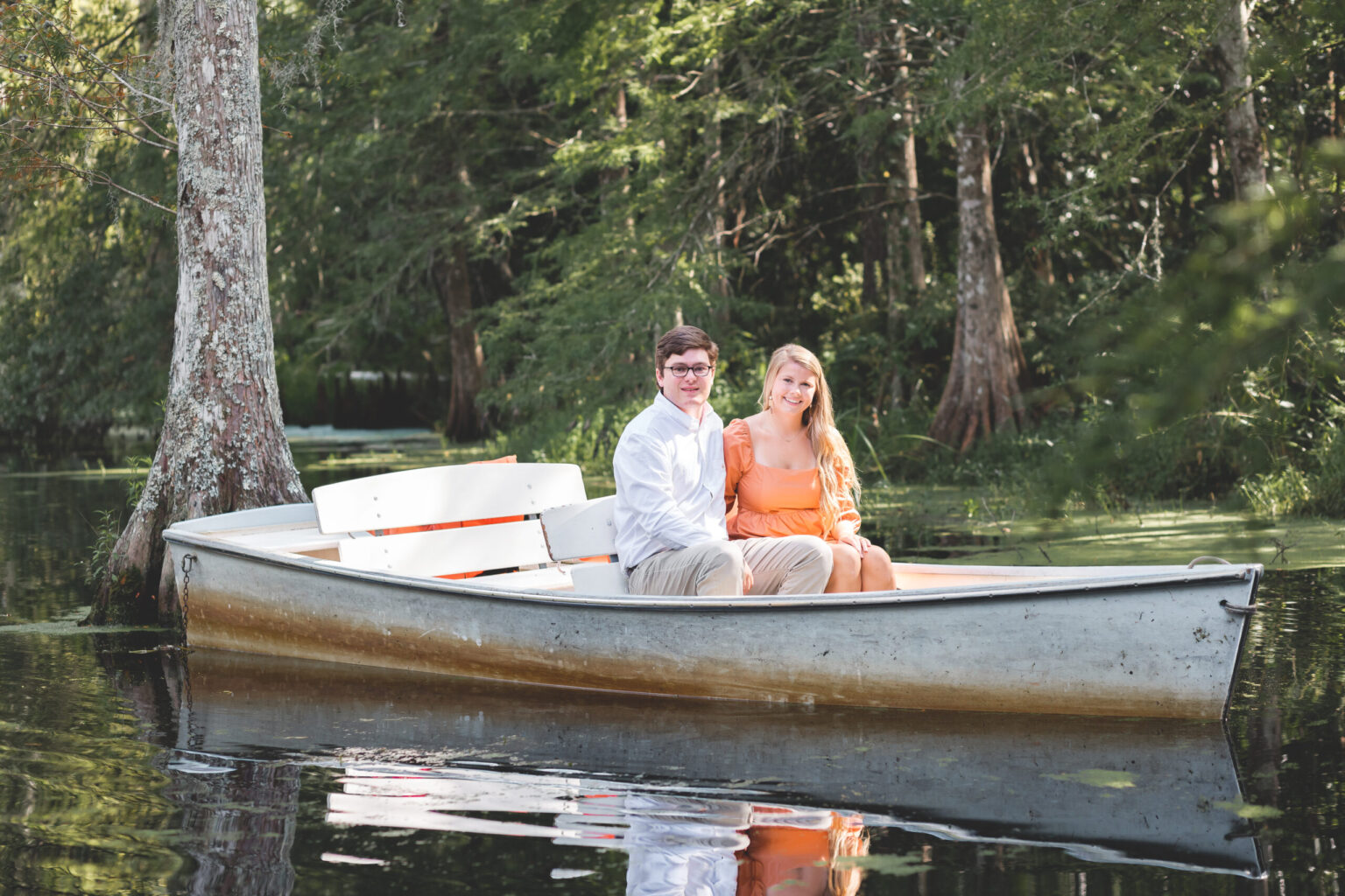 Elopement Wedding A man and woman sit together in a small boat on a calm, tree-lined lake. The man wears glasses, a white shirt, and khaki pants, while the woman is dressed in an orange dress. They are smiling and appear relaxed, surrounded by green trees and serene water. The boat has white seats and paddles. Elopements Inc