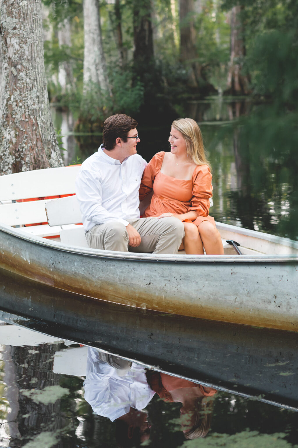 Elopement Wedding A man and woman sit in a small rowboat on a calm, reflective lake surrounded by trees. The man wears glasses, a white shirt, and khaki pants, while the woman wears an orange dress. They are smiling at each other, appearing happy and relaxed, with lush greenery in the background. Elopements Inc