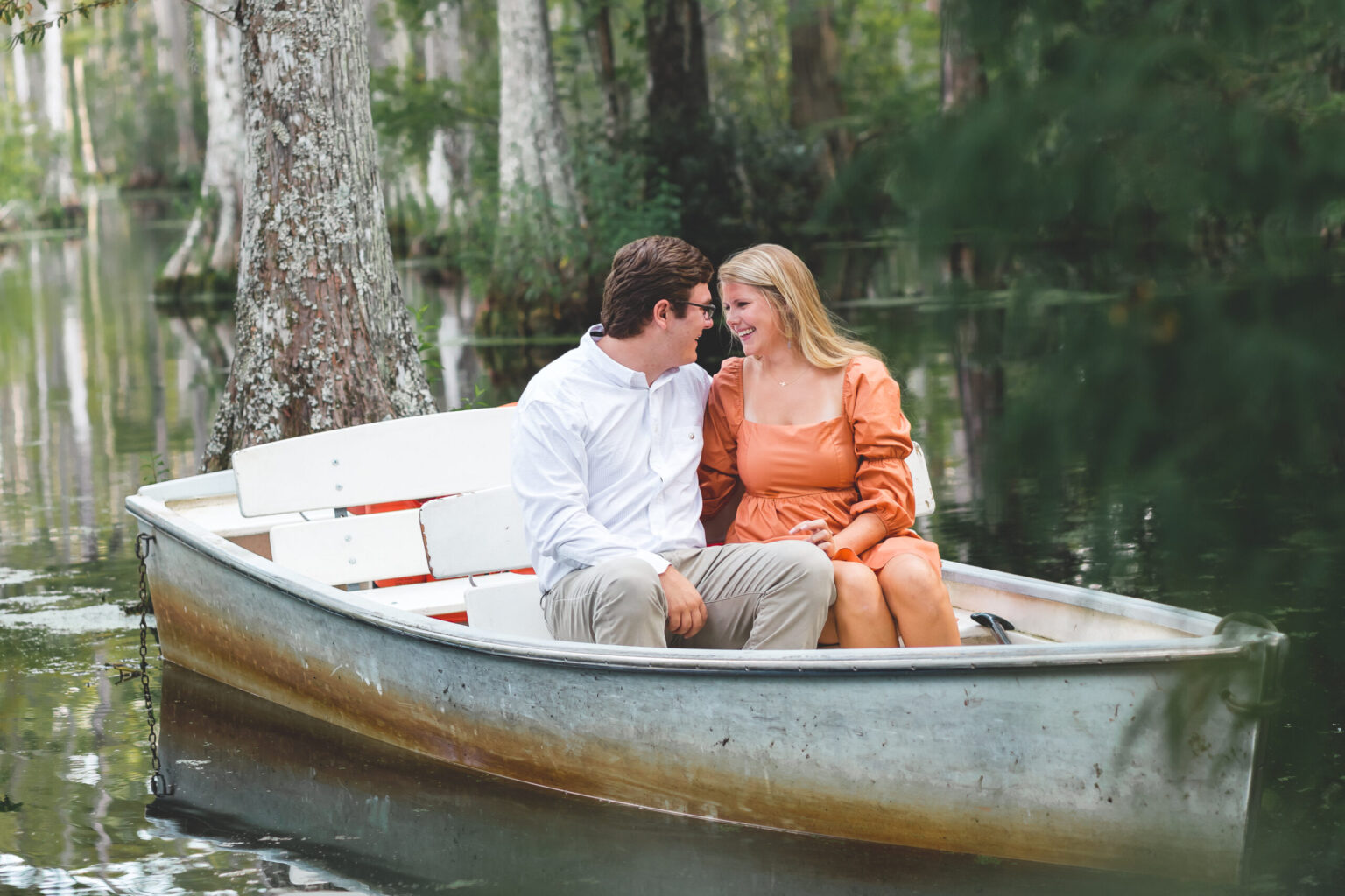 Elopement Wedding A couple sits close together in a small boat on a calm lake surrounded by trees. The man, dressed in a white shirt and khaki pants, and the woman in an orange dress, gaze at each other affectionately. The boat gently floats among tree trunks and greenery, creating a serene and romantic setting. Elopements Inc