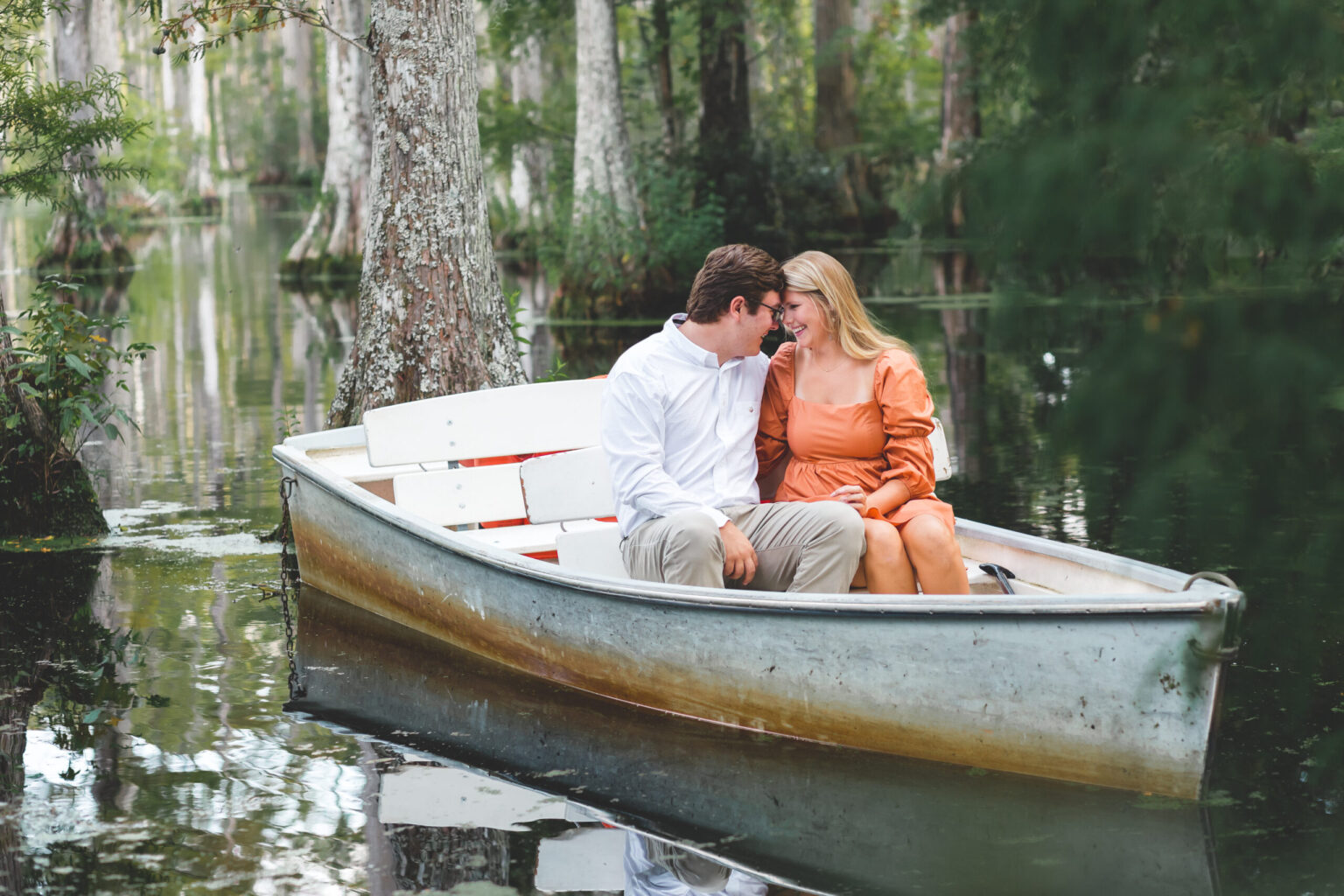 Elopement Wedding A couple sits in a small boat on a calm forested lake. The man wears a white shirt and light pants, while the woman wears an orange dress. They lean in close, smiling at each other surrounded by tall trees and lush greenery reflected in the still water. Elopements Inc