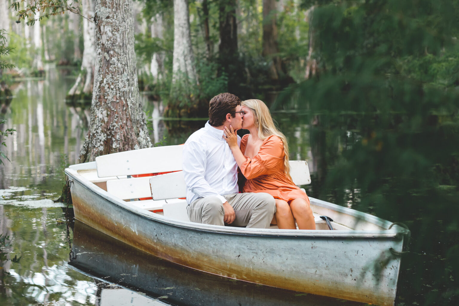 Elopement Wedding A couple sits in a rowboat on a serene forest pond, surrounded by trees and lush greenery. The man, in a white shirt and khaki pants, kisses the woman, who wears an orange dress. The scene is peaceful and intimate, with soft, natural light filtering through the tree canopy. Elopements Inc