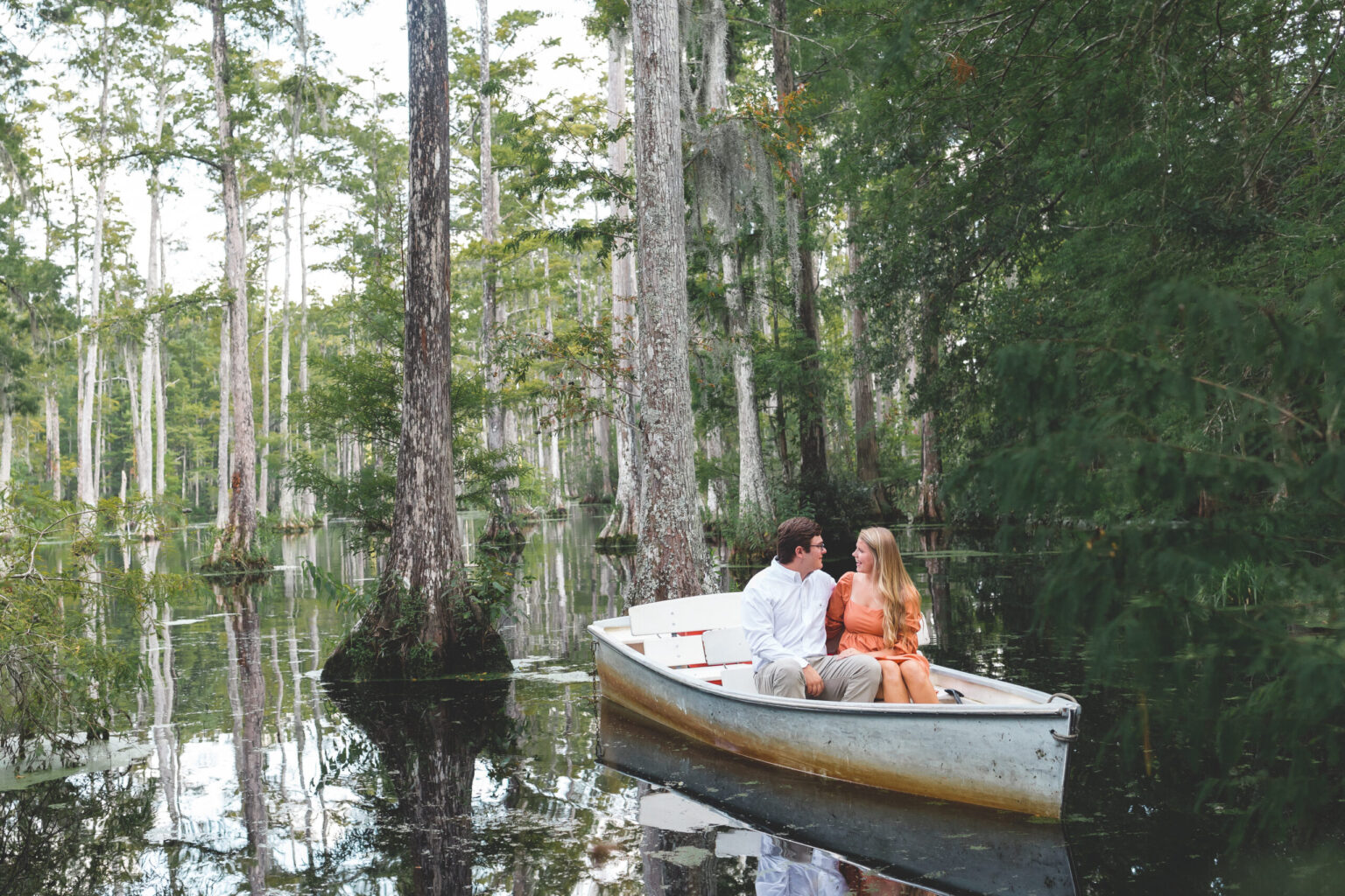 Elopement Wedding A couple is sitting in a small boat on a calm, reflective lake surrounded by tall trees and lush greenery. The man is wearing a white shirt and khaki shorts, while the woman is in an orange dress. They are looking at each other intimately, creating a romantic atmosphere in the serene setting. Elopements Inc