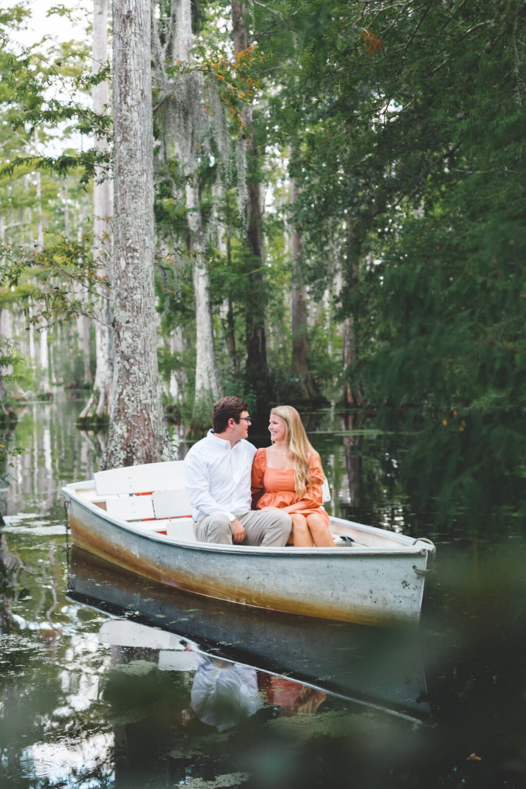 Elopement Wedding A couple is sitting in a small white rowboat on a serene, forested waterway. The man is wearing a white shirt and khaki pants, while the woman is in an orange dress. They are smiling and looking at each other, surrounded by tall trees and lush greenery reflected on the calm water. Elopements Inc