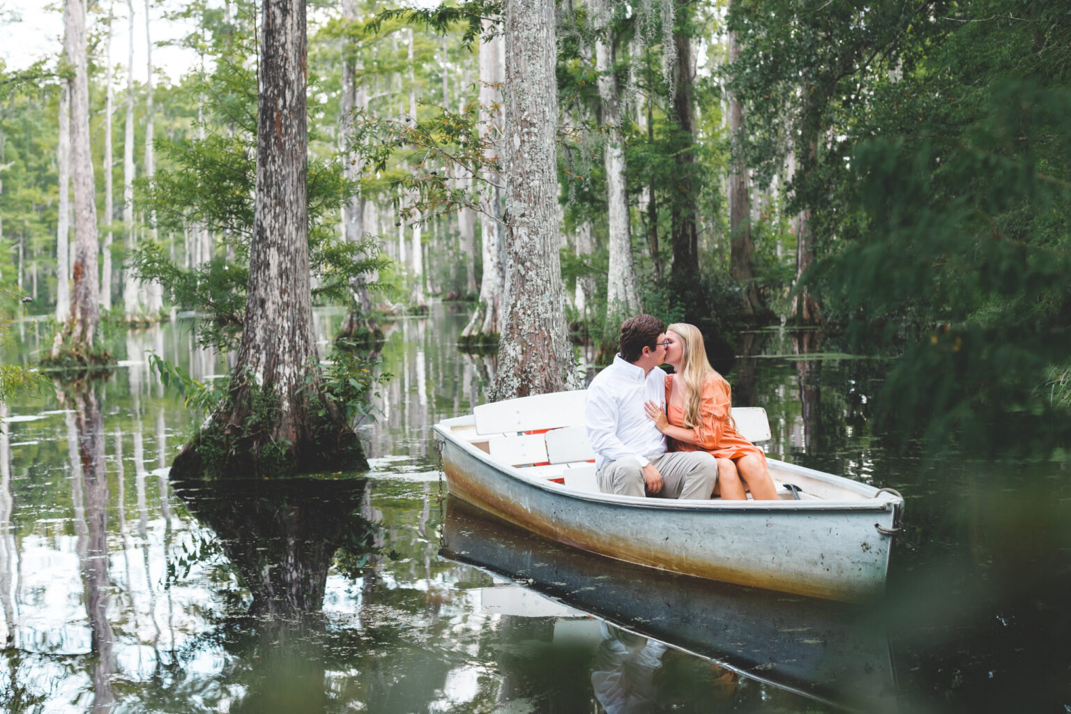 Elopement Wedding A couple sits in a rowboat on a serene lake surrounded by tall cypress trees. The man, wearing a white shirt, gently holds the woman, dressed in a flowing orange dress, as they share a kiss. The tranquil water reflects the lush greenery, creating a peaceful, romantic atmosphere. Elopements Inc