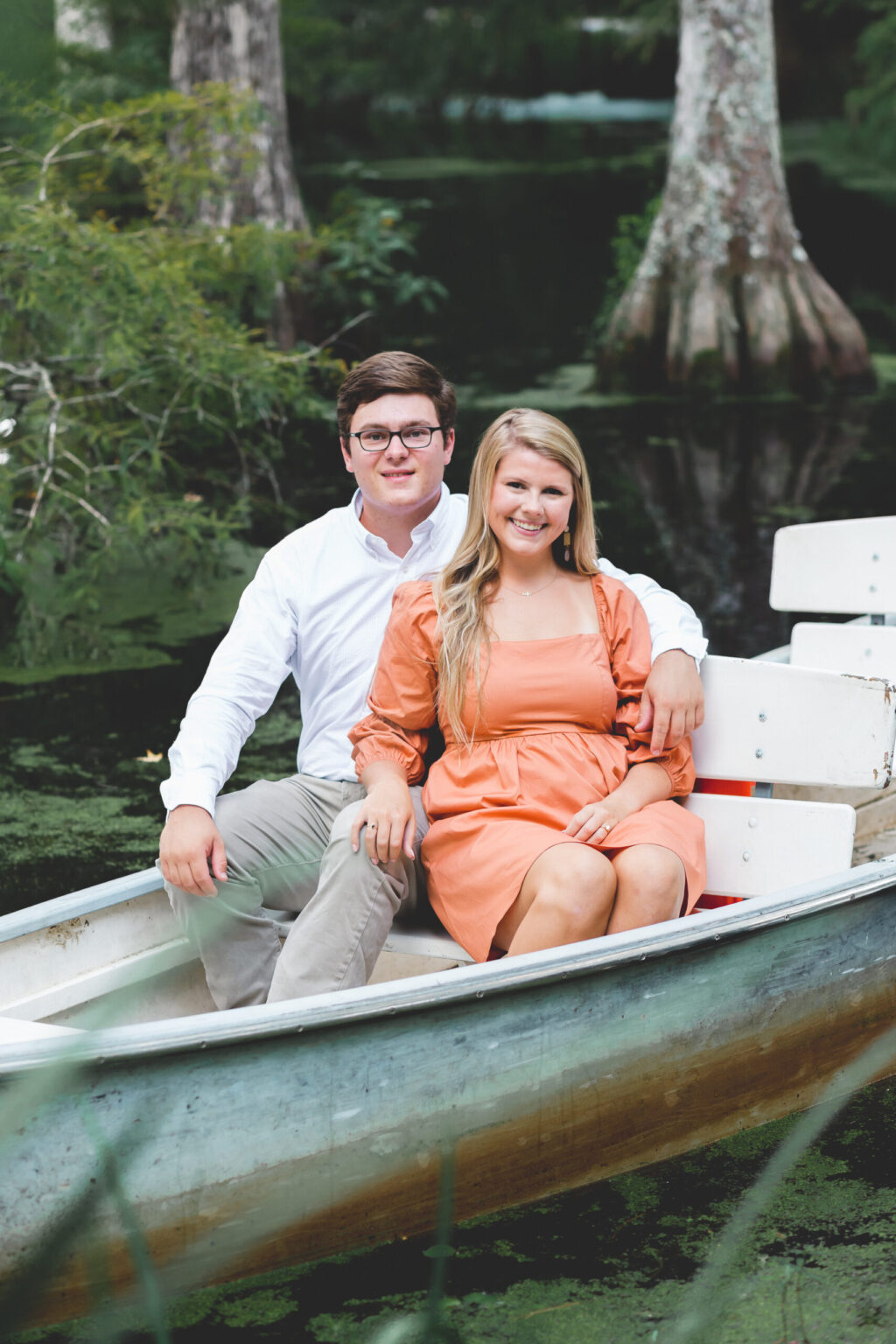 Elopement Wedding A couple sits together in a metal rowboat on a serene, tree-lined body of water. The man wears glasses, a white button-up shirt, and beige pants. The woman wears an orange dress and has long blonde hair. They are both smiling and appear relaxed, enjoying the peaceful surroundings. Elopements Inc