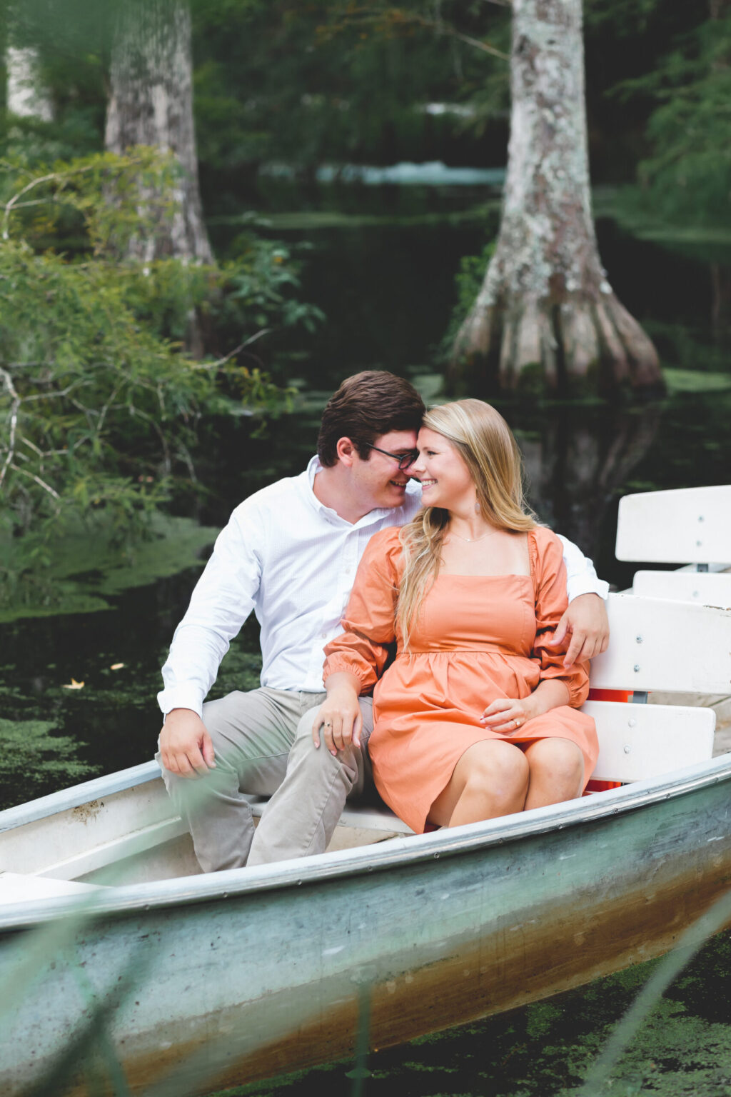 Elopement Wedding A couple sits in a boat on a tranquil water body surrounded by trees. The man, wearing a white shirt and beige pants, embraces a woman in an orange dress from behind. Both are smiling and gazing affectionately at each other. The scene is serene and romantic. Elopements Inc