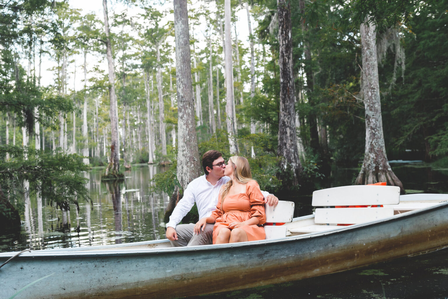 Elopement Wedding A couple sits in a rowboat on a serene, tree-lined lake. The man, wearing glasses and a white shirt, leans in to kiss the woman, who is in an orange dress. Tall trees with slender trunks surround the still water, creating a peaceful atmosphere. They look happy and relaxed, enjoying nature. Elopements Inc