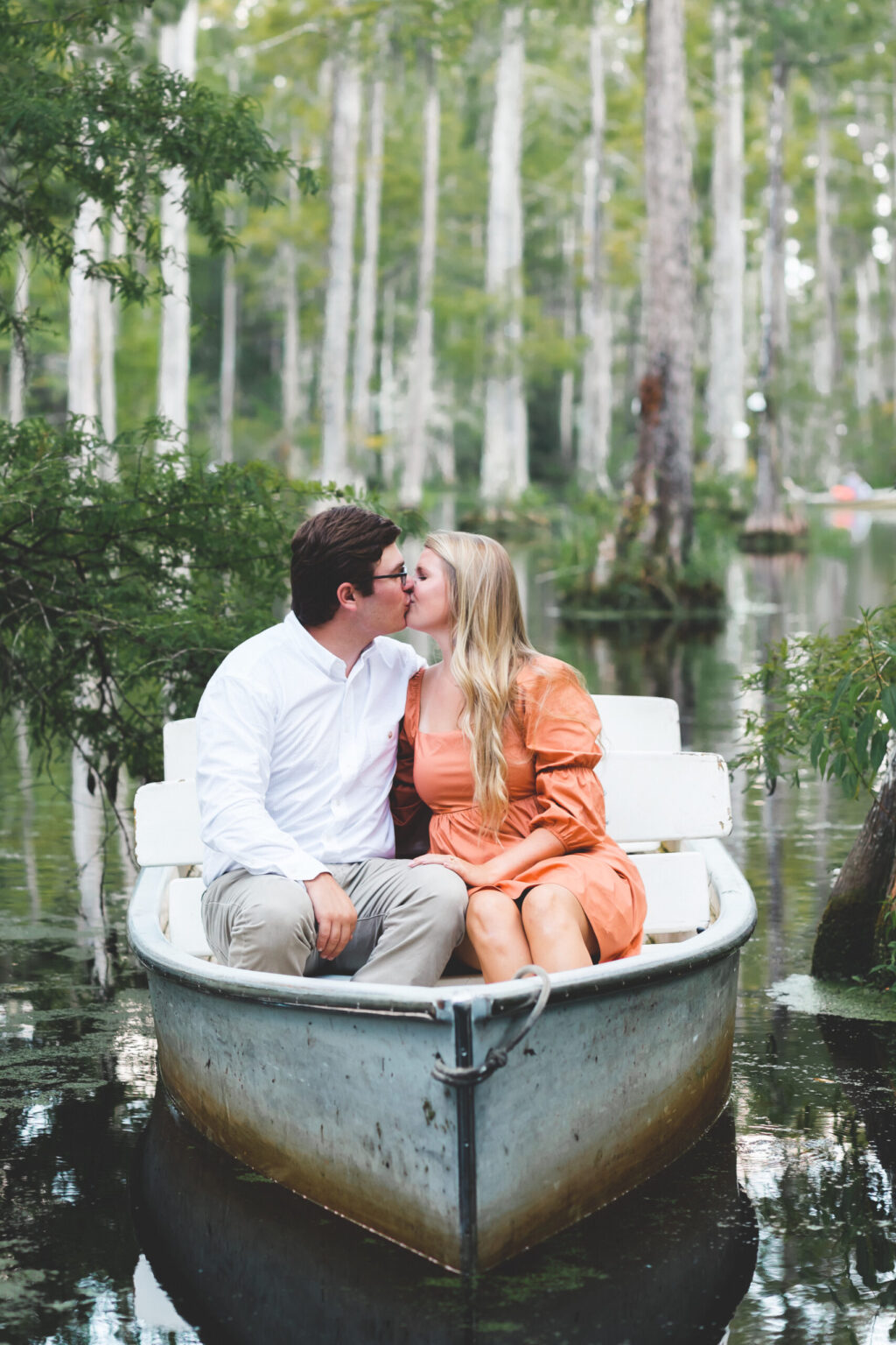 Elopement Wedding A couple shares a kiss while sitting in a small, white rowboat on a serene lake surrounded by lush greenery. The man wears a white shirt and beige pants, and the woman is in an orange dress. Tall trees with thin trunks and green foliage tower in the background, adding to the tranquil setting. Elopements Inc