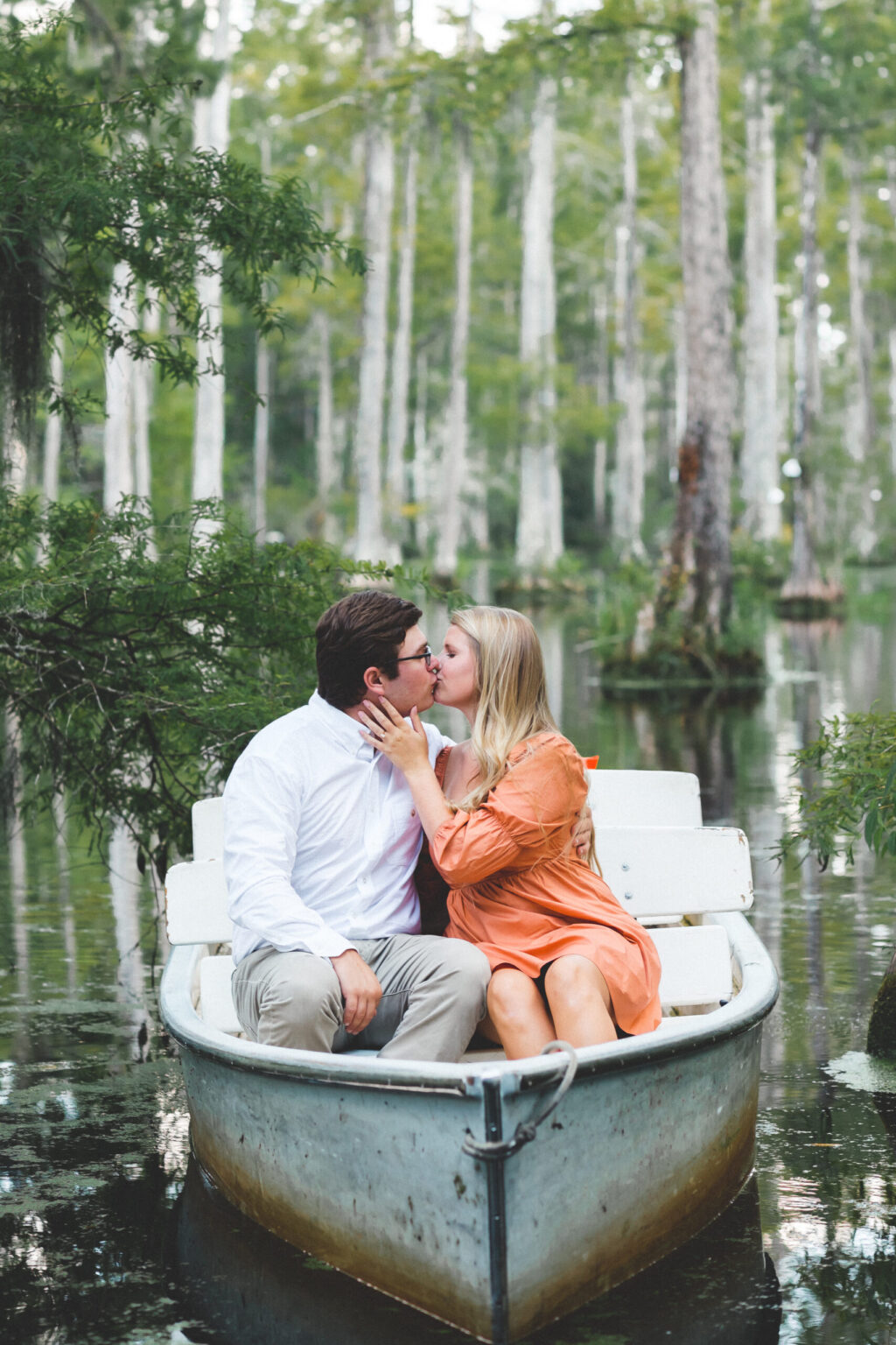 Elopement Wedding A couple sits in a small rowboat on a calm lake surrounded by lush greenery and tall trees. The man, dressed in a white shirt and beige pants, and the woman, wearing an orange dress, share a kiss. The serene natural setting includes reflections of the trees in the water. Elopements Inc