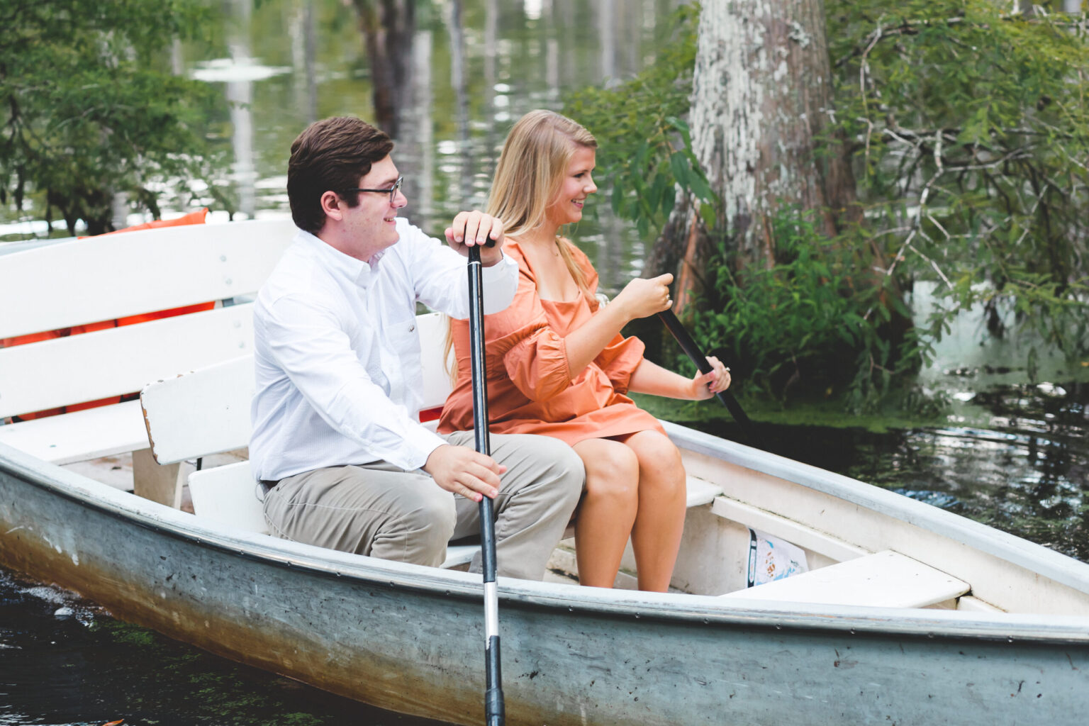 Elopement Wedding A man and woman are seated in a white boat on a calm body of water. The man, wearing a white shirt and glasses, is holding a paddle, while the woman, in an orange dress, also holds a paddle. They are surrounded by green trees and the water has reflections of the greenery. Elopements Inc