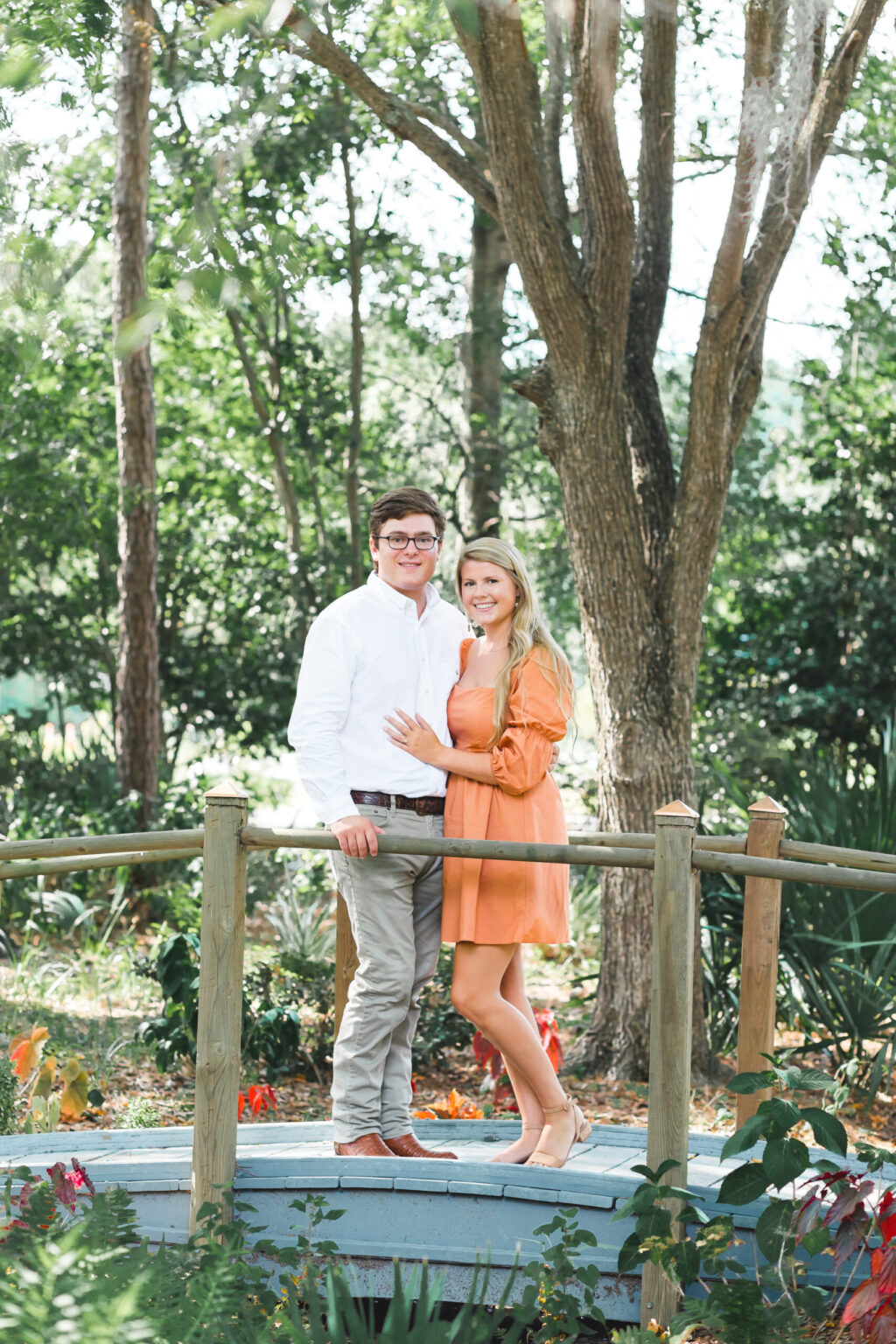 Elopement Wedding A couple stands closely together, smiling at the camera, on a small wooden bridge in a lush garden. The man wears a white shirt and khaki pants, while the woman wears an orange dress. Tall trees and greenery surround them, creating a serene and natural backdrop. Elopements Inc