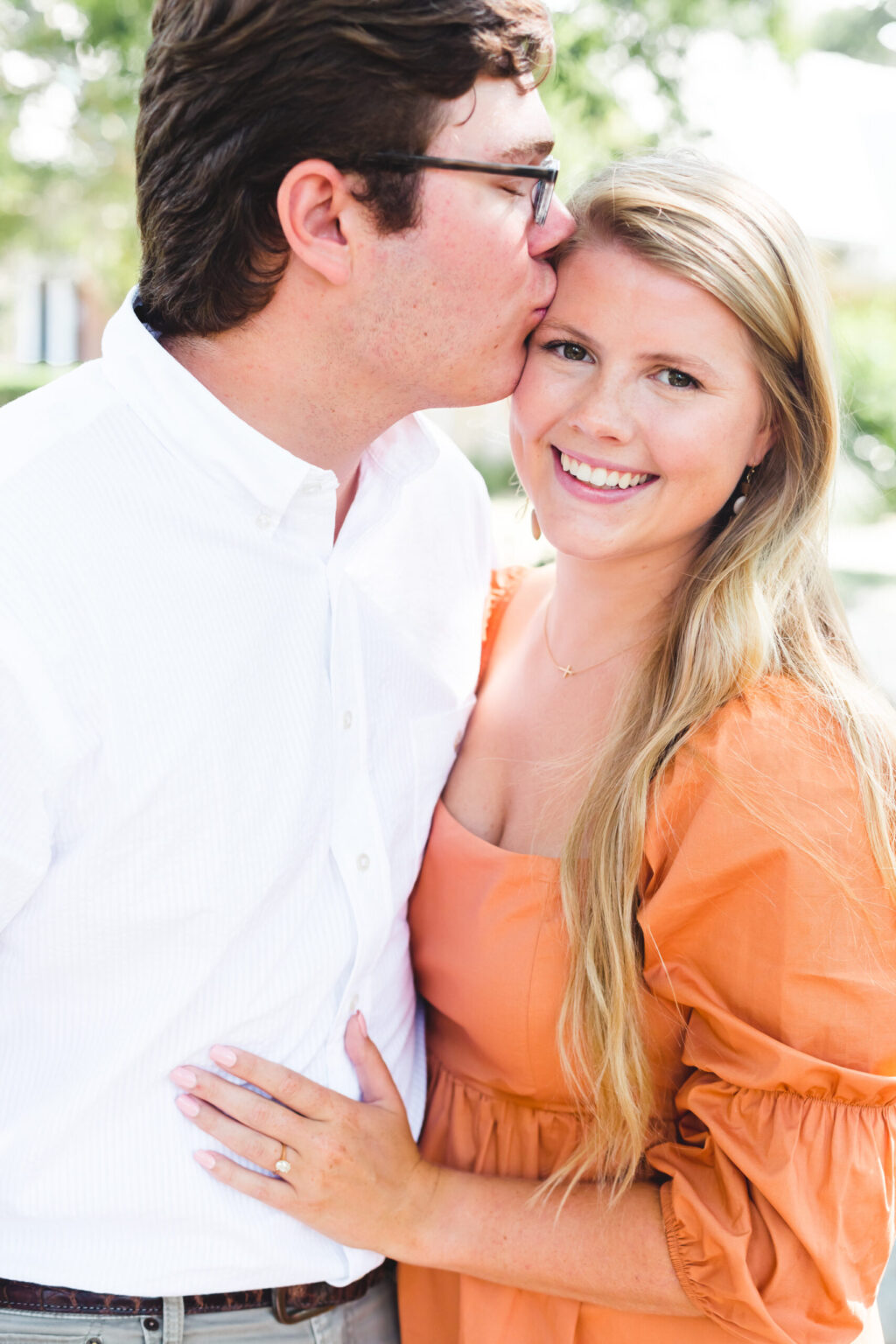 Elopement Wedding A man with short dark hair, glasses, and a white shirt kisses the forehead of a smiling woman with long blonde hair and an orange dress. They are standing outdoors, and the woman rests her hand, with a visible ring, on the man's chest. The background is blurred with greenery. Elopements Inc