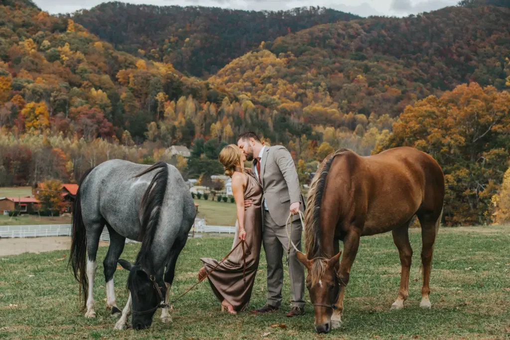 Elopement Wedding A couple dressed in formal attire stand closely together, sharing a kiss in an open grassy field with two horses grazing nearby. The backdrop features a scenic view of autumn-colored mountains and trees, creating a picturesque and romantic atmosphere. Elopements Inc