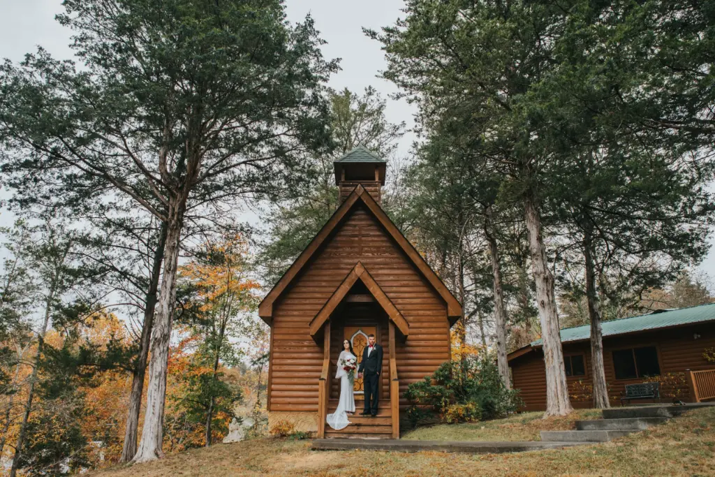 Elopement Wedding A newlywed couple stands at the entrance of a small, rustic, wooden chapel surrounded by tall trees with autumn foliage. The bride, in a white dress, holds a bouquet and the groom, in a suit, embraces her. The scene exudes a serene, natural ambiance with a mix of green and orange leaves. Elopements Inc