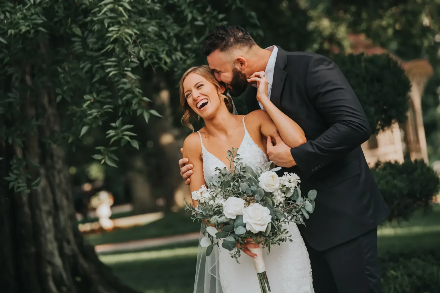 Elopement Wedding A bride and groom share a joyful moment outdoors. The bride, in a white lace wedding dress, holds a bouquet with white flowers and greenery, laughing as the groom in a dark suit playfully kisses her head. They stand under a lush tree, with soft sunlight filtering through the leaves. Elopements Inc