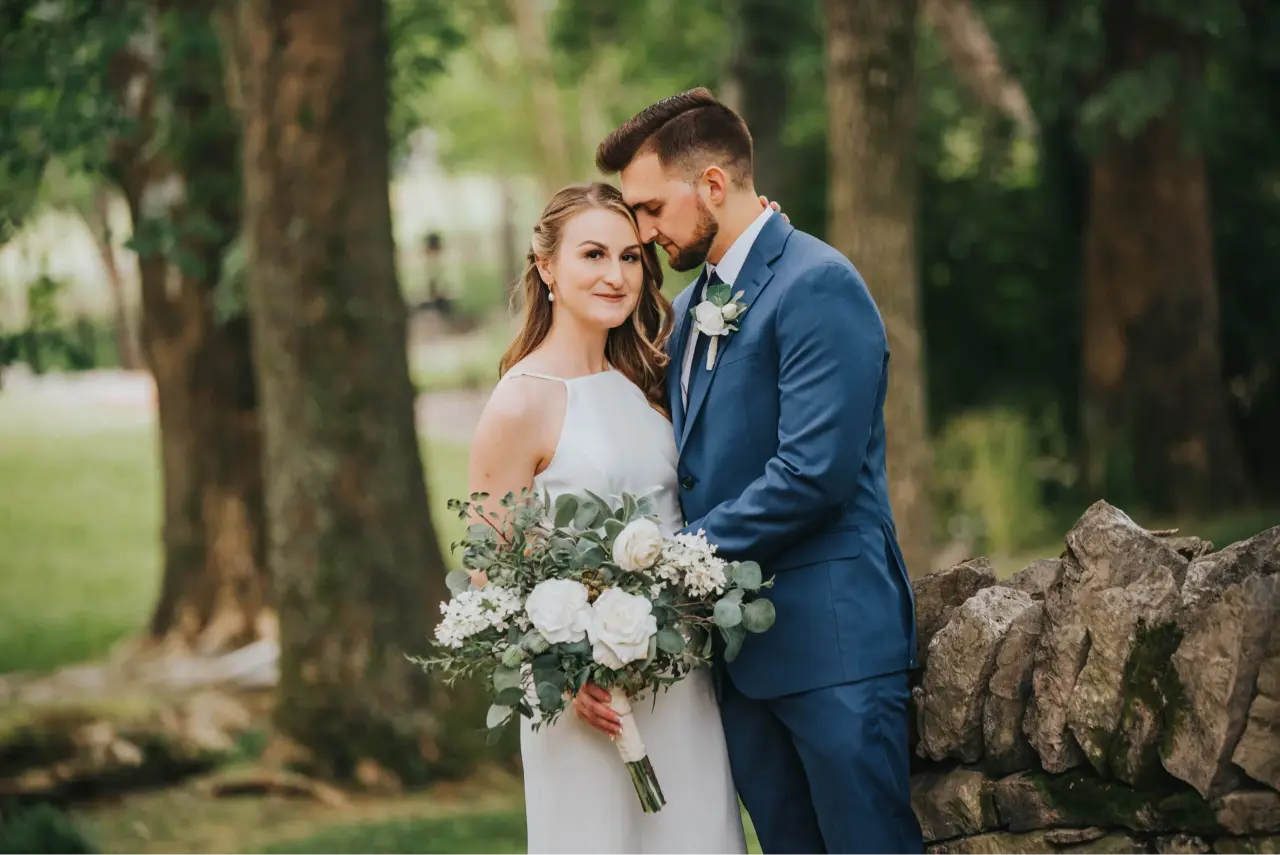 Elopement Wedding A bride in a white sleeveless gown holds a lush bouquet while standing beside a groom in a blue suit and tie. They stand close together, the groom gently leaning his forehead against the bride's temple. They are outdoors, surrounded by trees and greenery, near a rustic stone wall. Elopements Inc