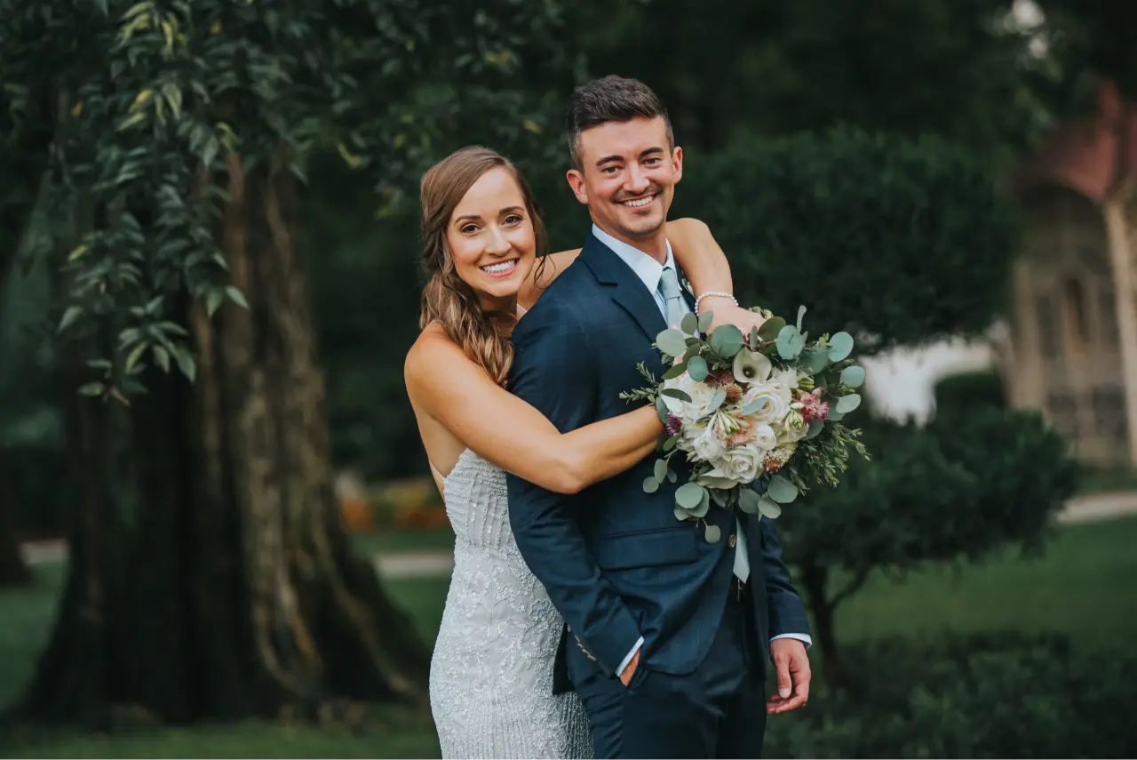 Elopement Wedding A bride in a white, strapless, sequined gown stands behind a groom in a dark blue suit, smiling and holding a bouquet of flowers. She has one arm around his shoulders, and they are standing outdoors in a park-like setting with green trees and bushes in the background. Elopements Inc