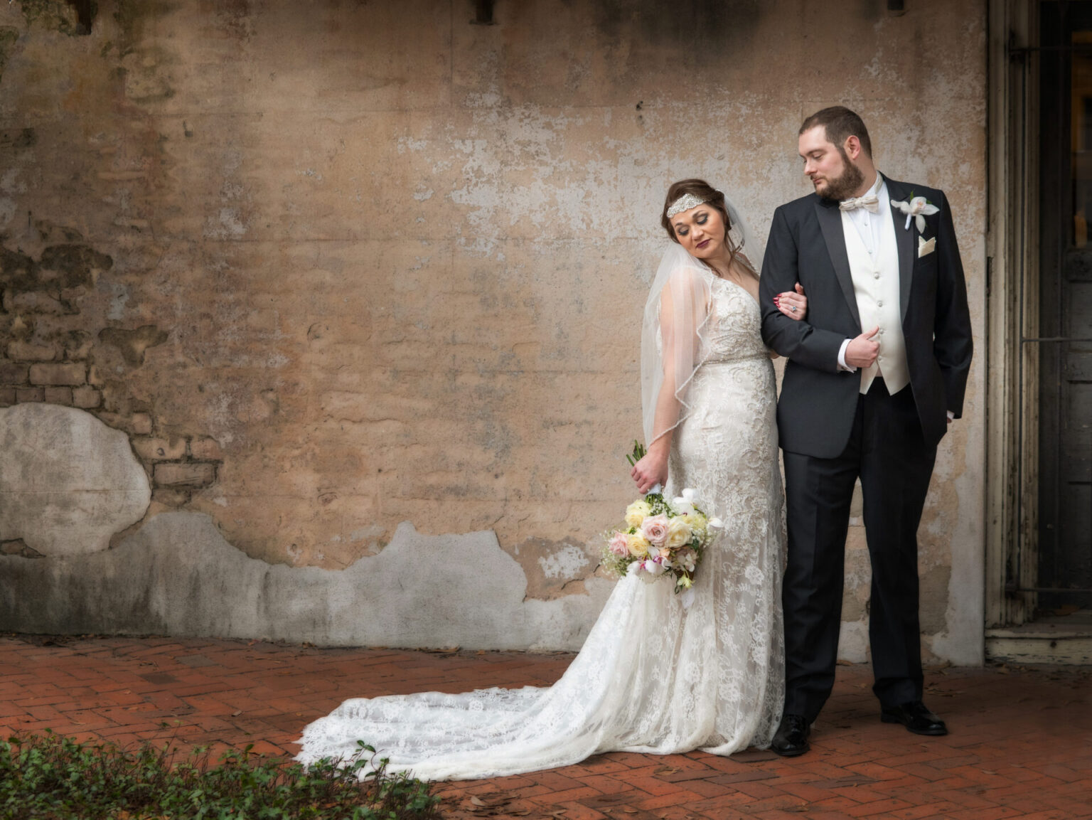 Elopement Wedding A bride and groom stand together against a rustic, textured wall. The bride wears a lace wedding gown with a veil and holds a bouquet of pink and white flowers. The groom is in a black suit with a white shirt and tie. They look at each other, exuding a romantic and elegant atmosphere. Elopements Inc