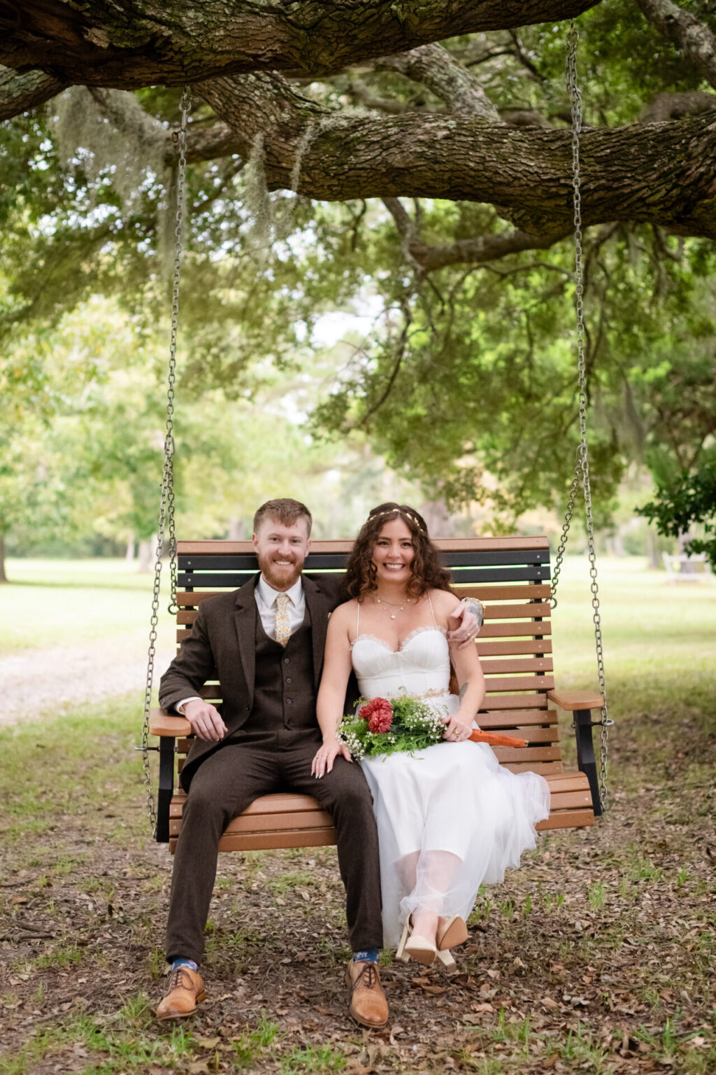 Elopement Wedding A couple sits on a wooden swing under a large tree. The woman is in a white dress holding a bouquet, and the man wears a brown suit. They are smiling, surrounded by green foliage and a grassy field. The scene conveys a romantic, outdoor atmosphere. Elopements Inc