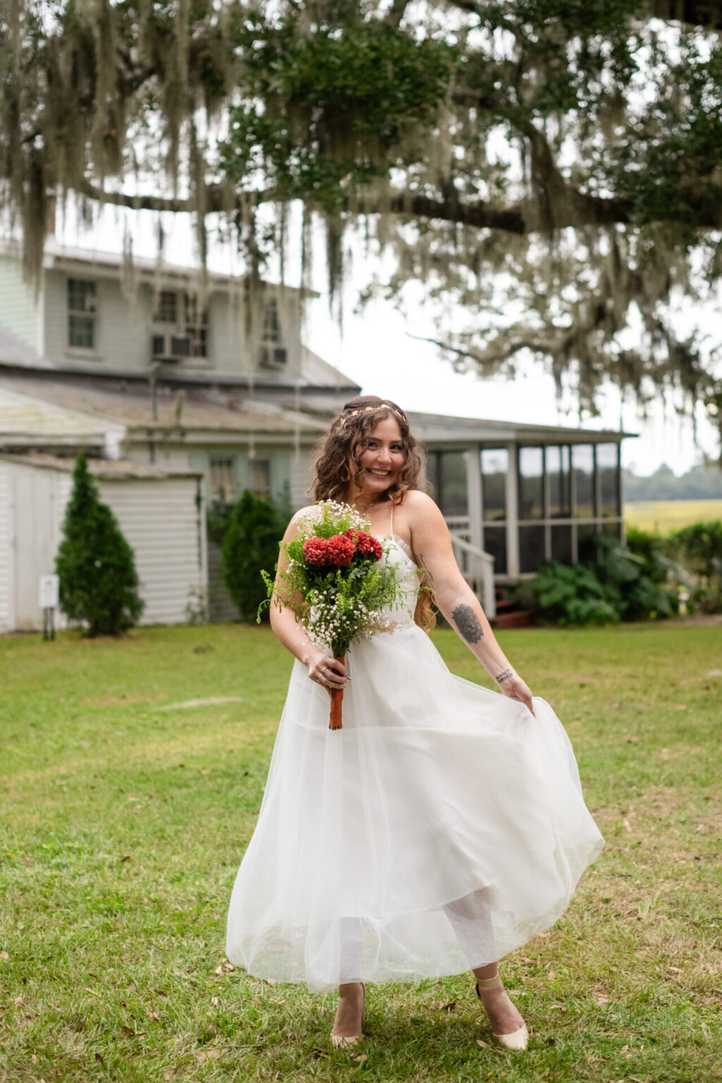 Elopement Wedding A bride in a flowing white dress smiles and holds a bouquet of red and green flowers. She stands on grass with trees and a rustic house in the background. Her curly hair is styled loosely, and she is playfully twirling her dress outside. Elopements Inc