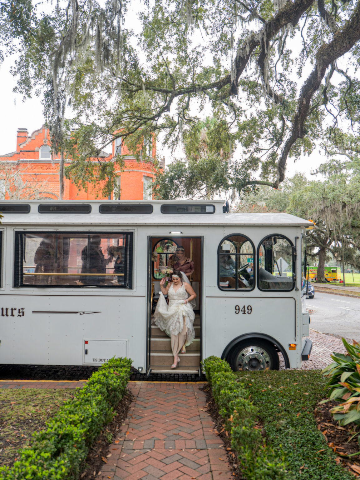 Elopement Wedding A bride and groom step off a white trolley, surrounded by lush greenery and brick pathways. The trolley displays the number "949." In the background, there's a red brick building and large trees draped with Spanish moss, creating a picturesque setting. Elopements Inc