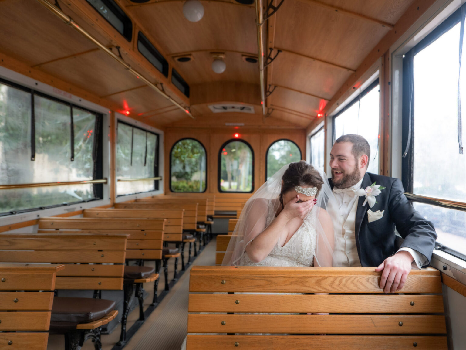 Elopement Wedding A bride and groom sitting on a wooden seat inside a vintage trolley. The bride, wearing a white dress and veil, covers her face with one hand while laughing. The groom, in a dark suit with a white boutonniere, smiles at her. The trolley is empty, featuring wooden bench seating. Elopements Inc