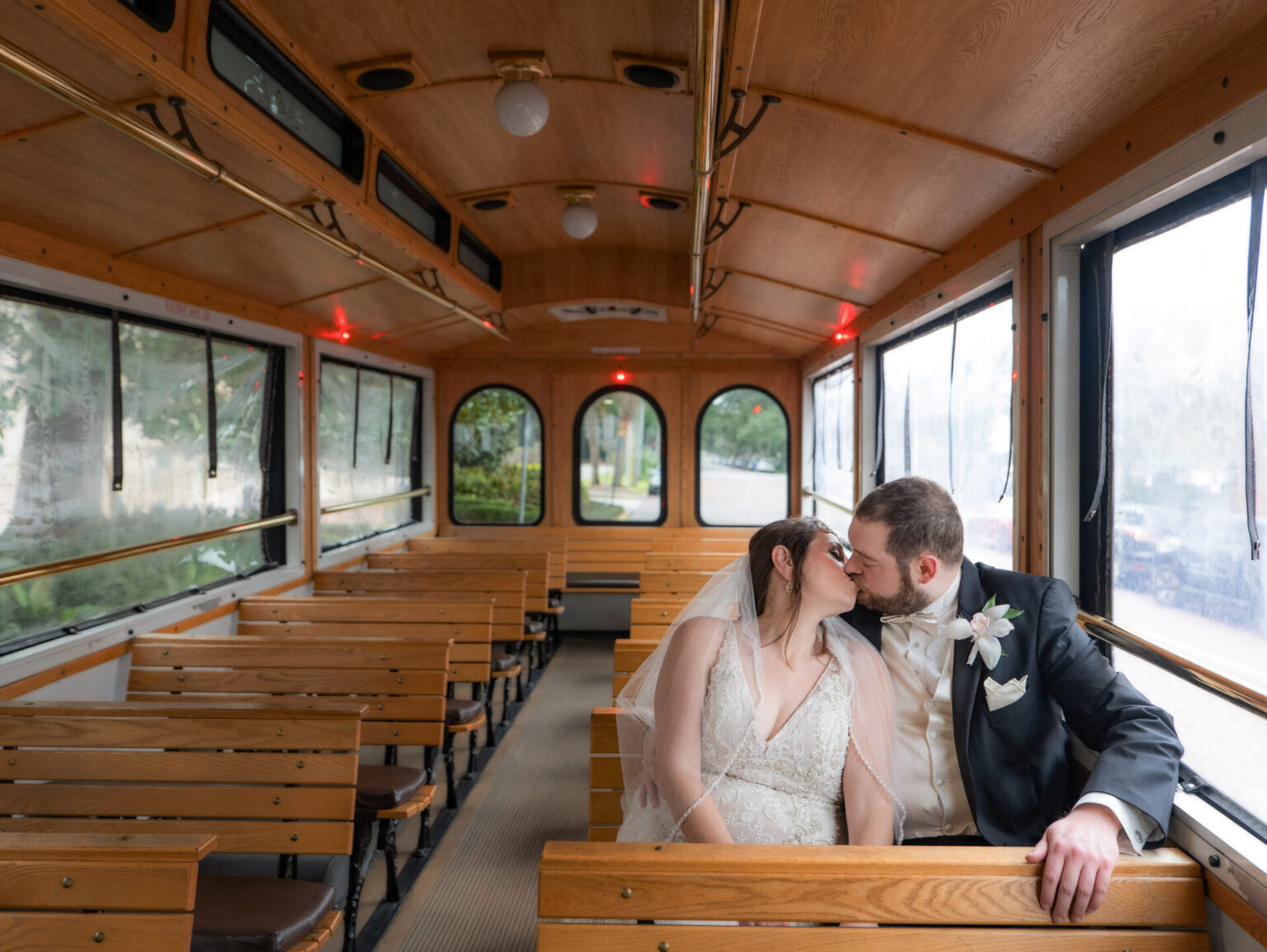 Elopement Wedding A bride and groom kiss while sitting together on a wooden trolley. The bride wears a white dress and veil, and the groom is in a dark suit with a boutonniere. The trolley is empty except for them, and soft light filters through the windows. Elopements Inc