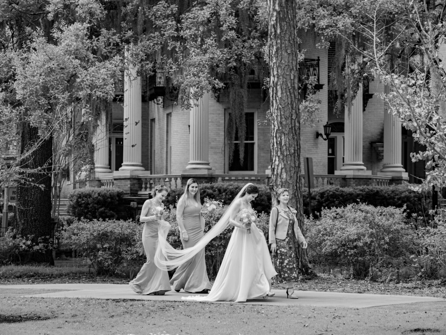 Elopement Wedding A black and white photo shows a bride and three women walking on a path lined with trees and bushes. The bride wears a long gown and veil, holding a bouquet. The other women wear elegant dresses. They approach a large building with tall columns in the background. Elopements Inc