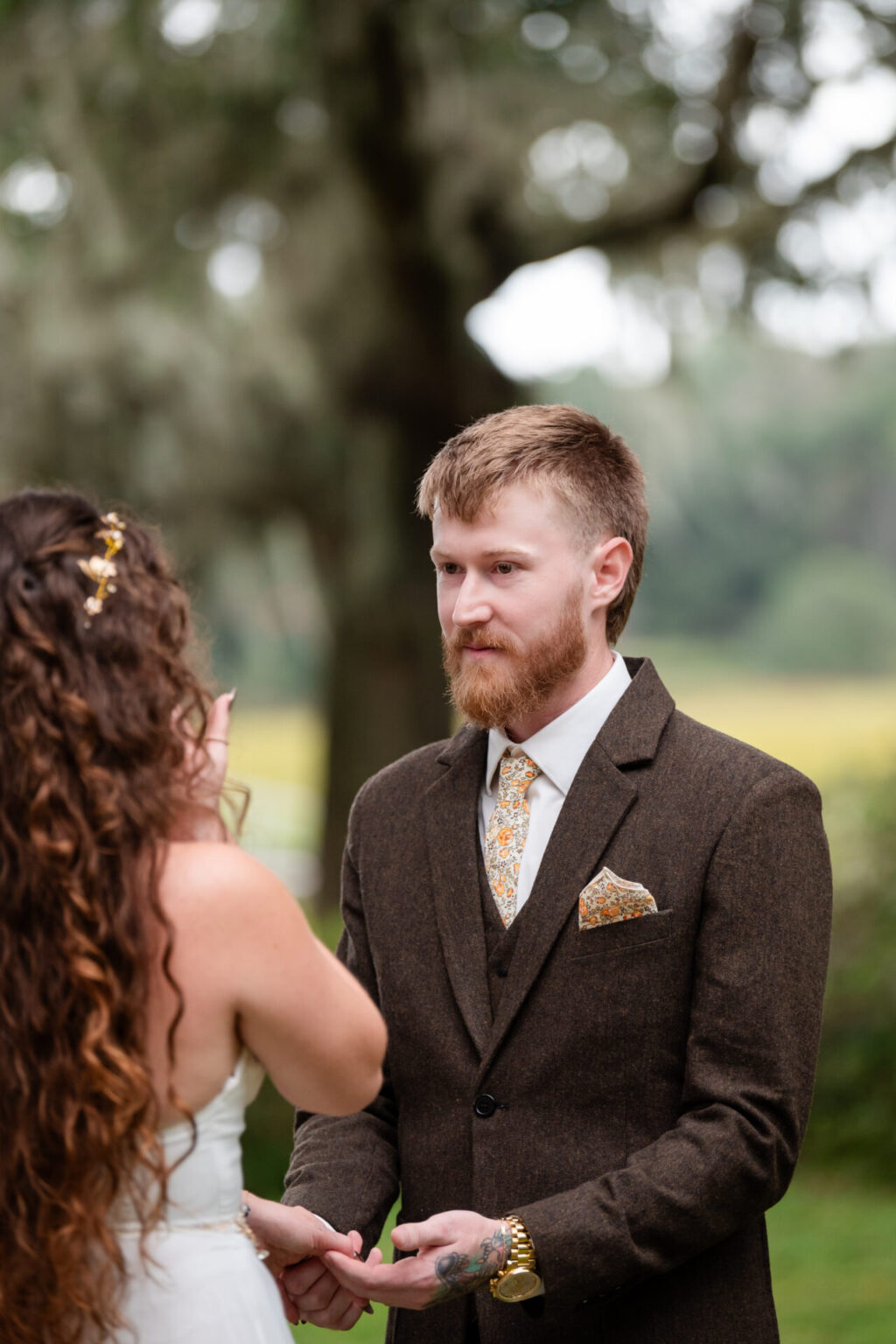 Elopement Wedding A groom with a short beard, wearing a brown suit and floral tie, stands outdoors, facing a bride with long curly hair. The bride is wearing a white gown with gold hair accents. They are holding hands under a tree with soft-focus greenery in the background. Elopements Inc