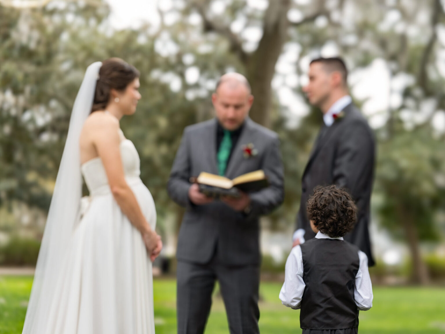 Elopement Wedding A bride in a white dress and veil stands beside a groom in a dark suit. An officiant in a gray suit holds an open book. A young boy, wearing a black vest and white shirt, faces them. They're outdoors, with blurred greenery in the background, suggesting a wedding ceremony. Elopements Inc