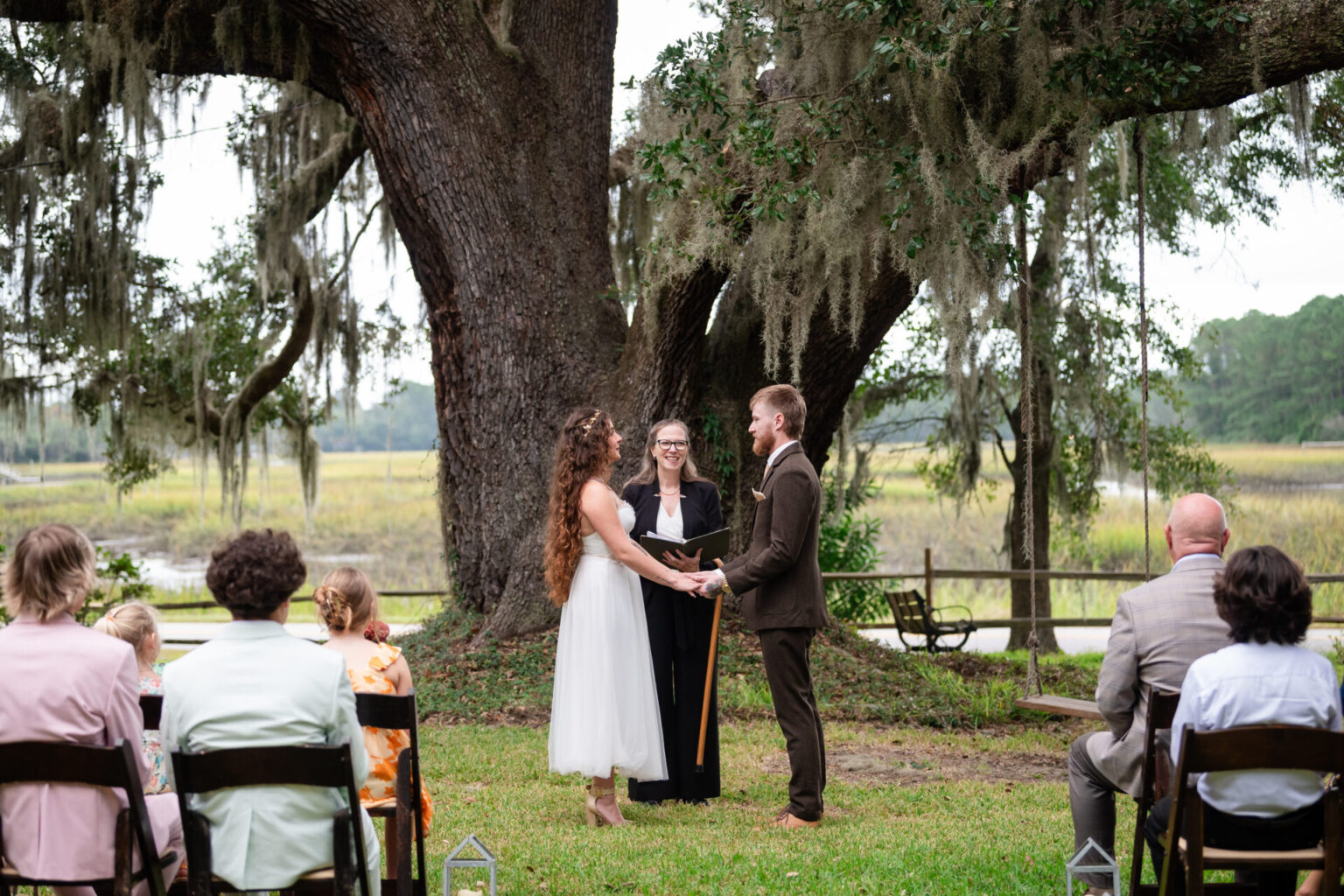 Elopement Wedding A bride and groom stand holding hands during an outdoor wedding ceremony under a large, sprawling oak tree draped with Spanish moss. They face an officiant. Guests sit on wooden chairs in two rows, observing the scene. The background features lush greenery and a serene waterway. Elopements Inc