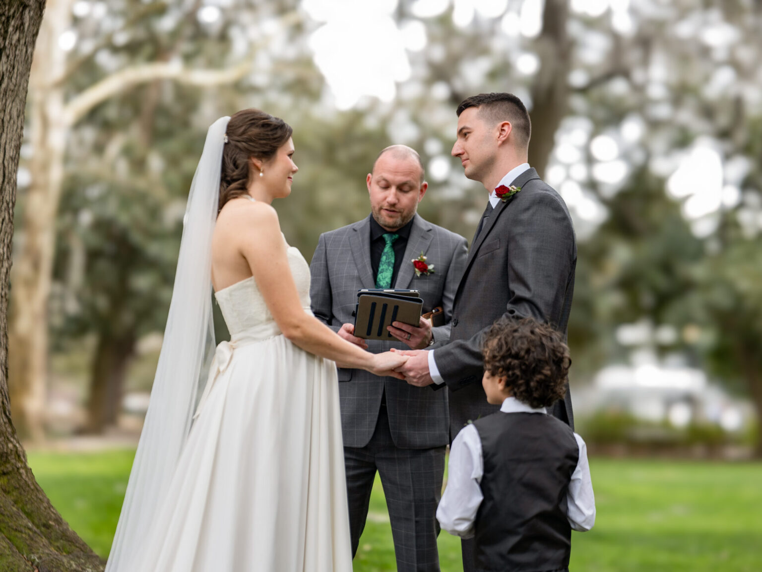 Elopement Wedding A bride and groom stand facing each other, holding hands, during an outdoor wedding ceremony. The officiant, wearing a suit, holds a tablet. A young boy in a vest stands near the groom. Trees and grass provide a natural backdrop, creating a serene and intimate setting. Elopements Inc