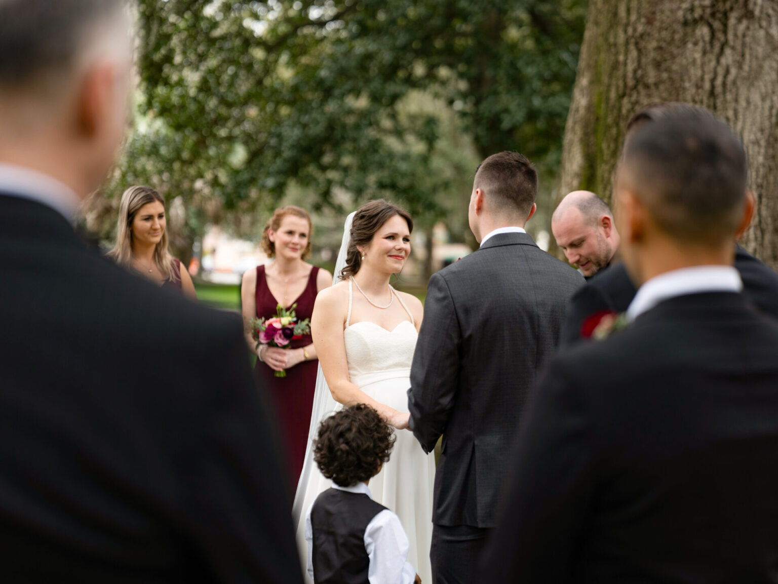 Elopement Wedding A bride in a white dress smiles while facing the groom during an outdoor wedding ceremony. Bridesmaids in burgundy dresses stand nearby holding flowers. A young boy in a vest is in the foreground. The scene is set in a park with large trees and greenery. Elopements Inc