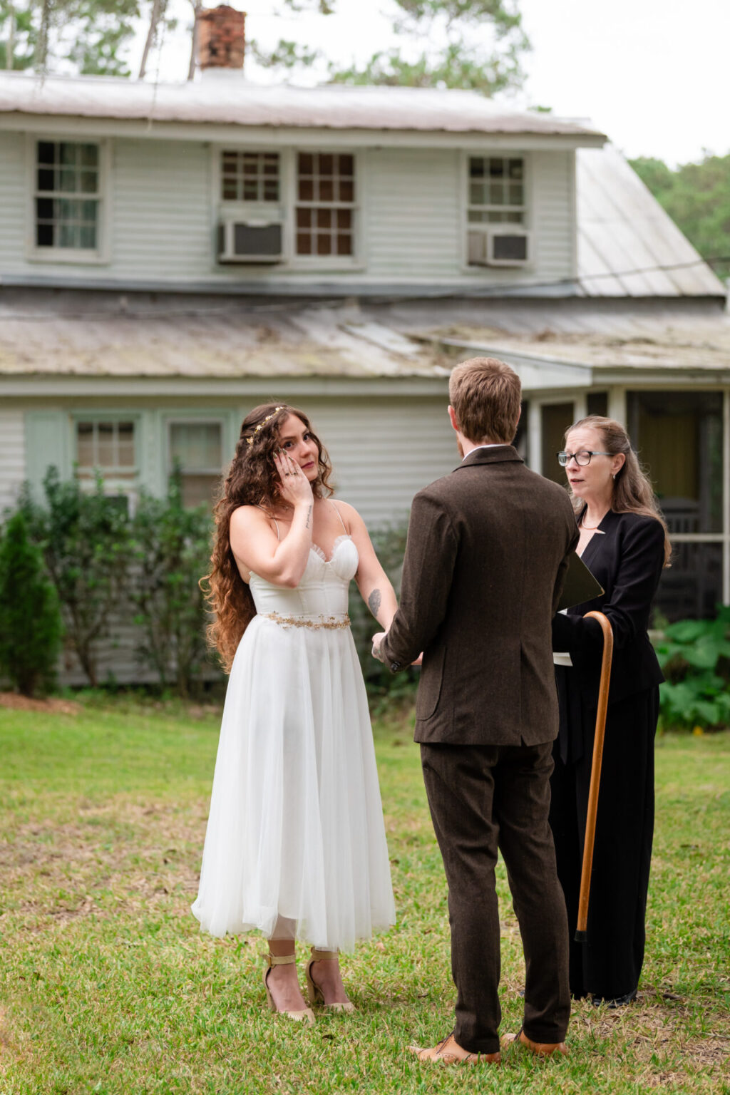 Elopement Wedding A bride in a white dress and a groom in a dark suit stand holding hands during a wedding ceremony outdoors. An officiant with glasses and a book stands beside them. The background features a two-story white house with a tin roof and greenery around the lawn. Elopements Inc