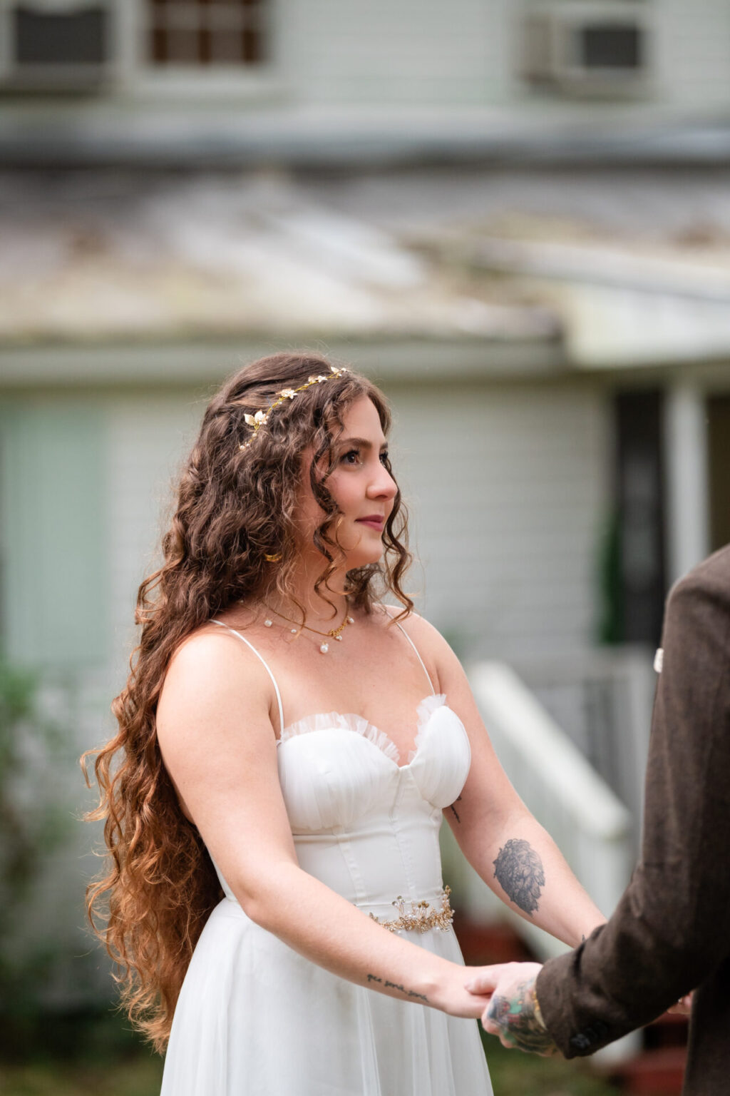 Elopement Wedding A bride with long, curly hair adorned with a floral headpiece and wearing a white wedding dress stands outdoors, holding hands with another person. She has a tattoo on her forearm. The background features a house with cream-colored siding and a sloping roof. Elopements Inc