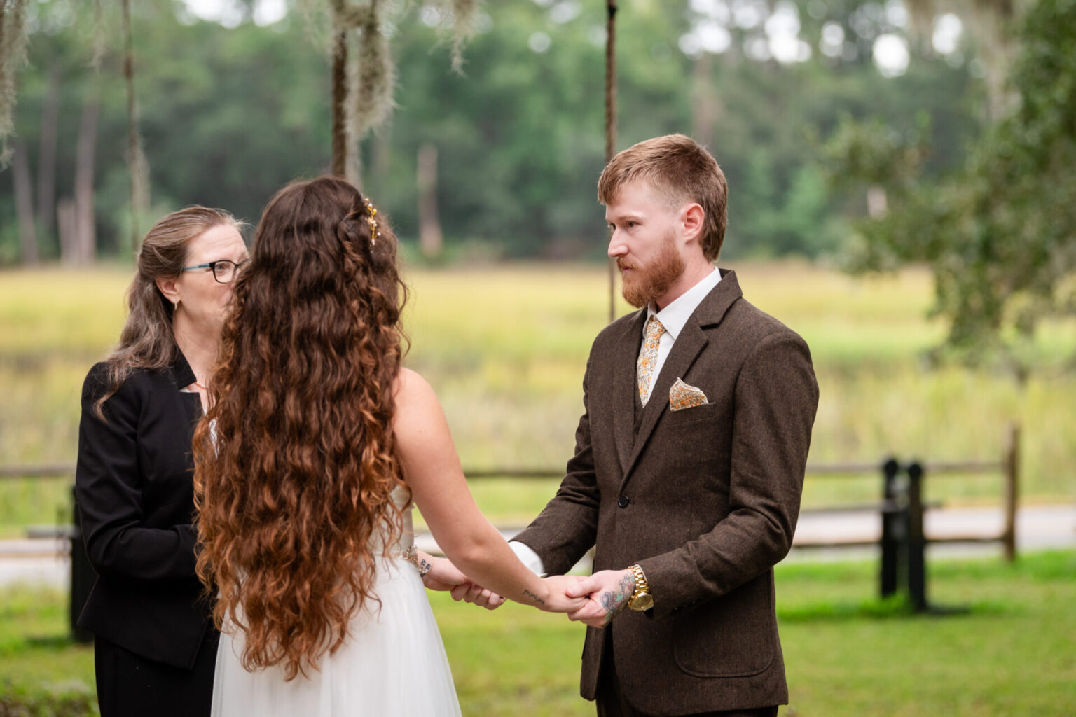 Elopement Wedding A couple holds hands during an outdoor wedding ceremony. The bride, with long, wavy hair, wears a white dress. The groom, with a beard, wears a brown suit and patterned tie. An officiant stands beside them. The background features greenery, trees, and a rustic wooden fence. Elopements Inc