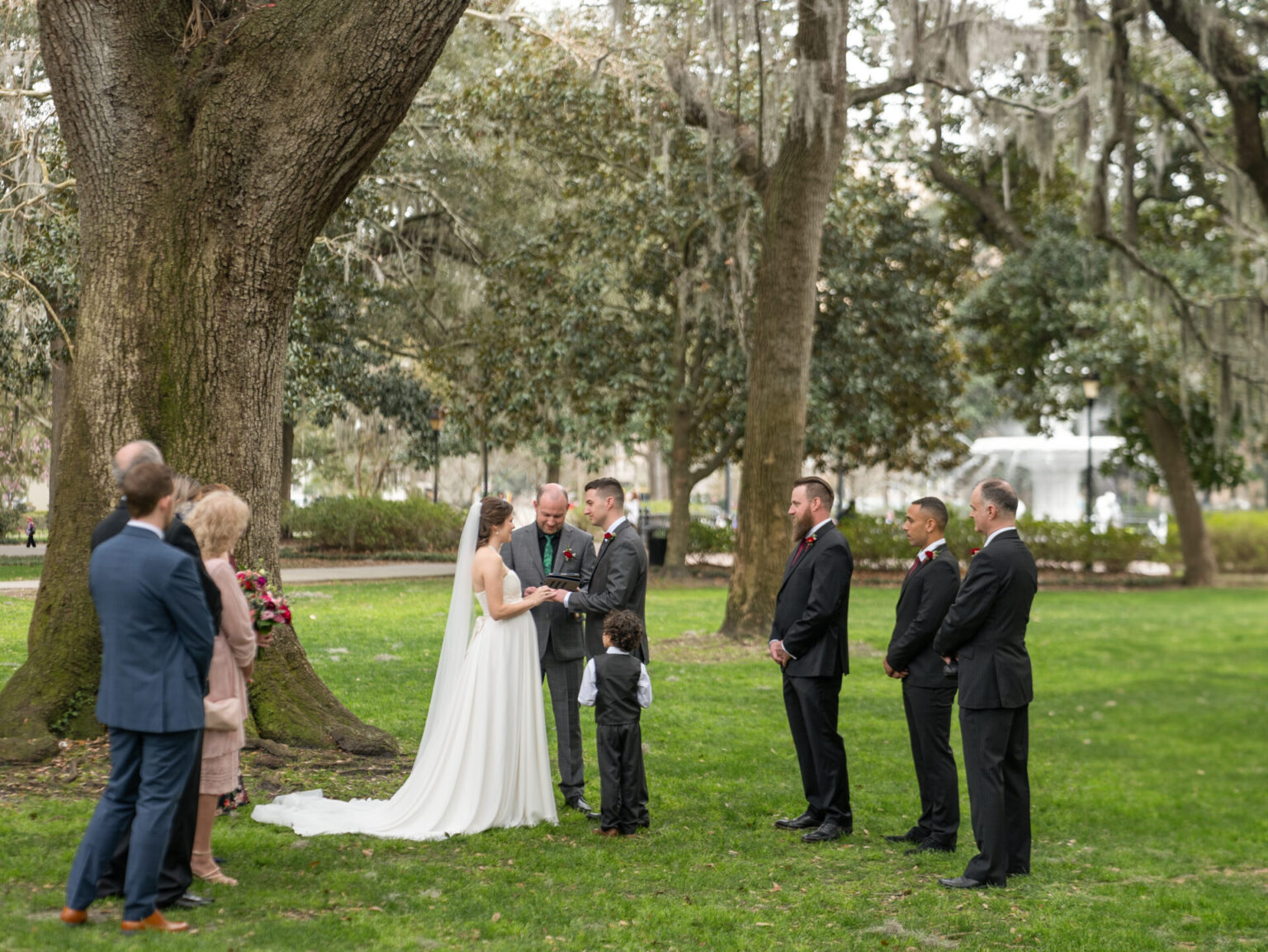 Elopement Wedding A couple stands under a large tree exchanging vows during an outdoor wedding ceremony. The bride wears a white dress, and the groom wears a gray suit. There are six guests; four men in suits, a woman in pink, and a child. The setting is a park with lush greenery and moss-draped trees. Elopements Inc