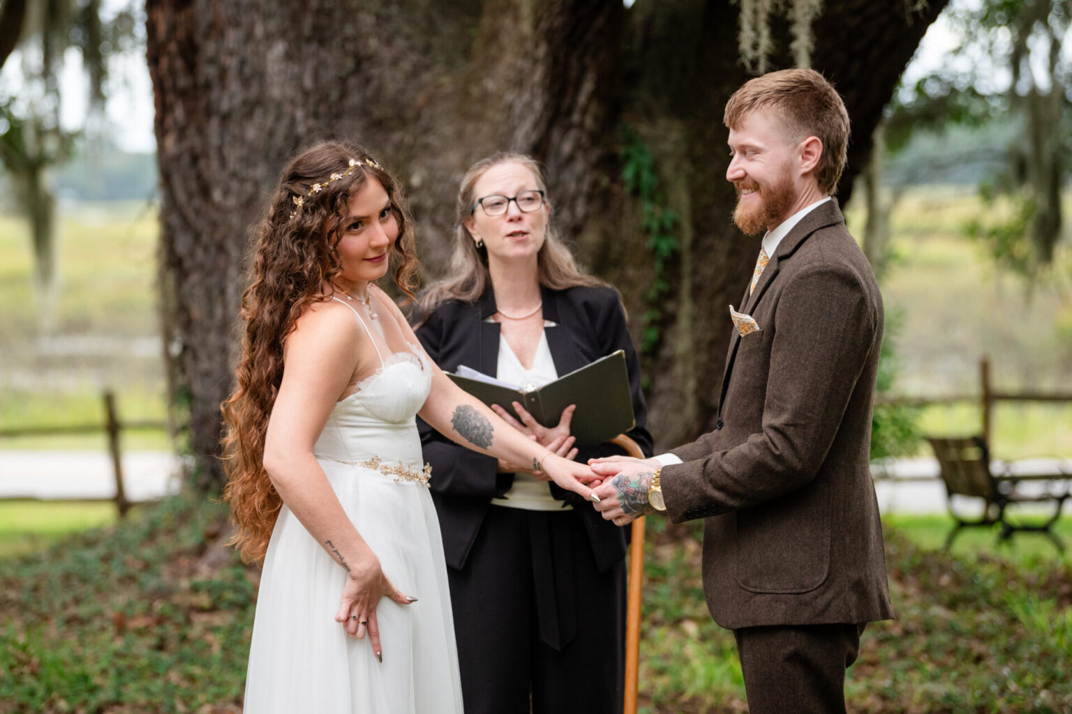 Elopement Wedding A couple is getting married outdoors under a large tree. The bride, in a white dress and gold headpiece, holds hands with the groom, who is wearing a brown suit. Both smile as an officiant, dressed in black and holding an open book, conducts the ceremony. Rustic scenery surrounds them. Elopements Inc