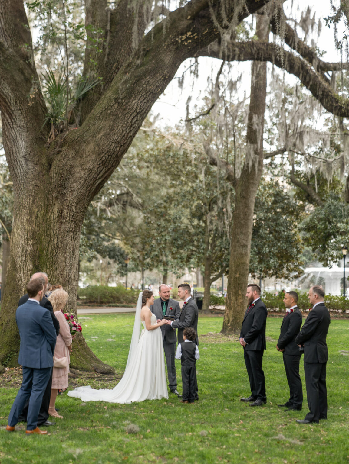 Elopement Wedding A wedding ceremony in a park setting under a large, moss-covered tree. The bride and groom stand facing the officiant, with the bride in a white dress and veil. Guests, including children dressed formally, are gathered around, observing. The grass is lush, and trees fill the background. Elopements Inc