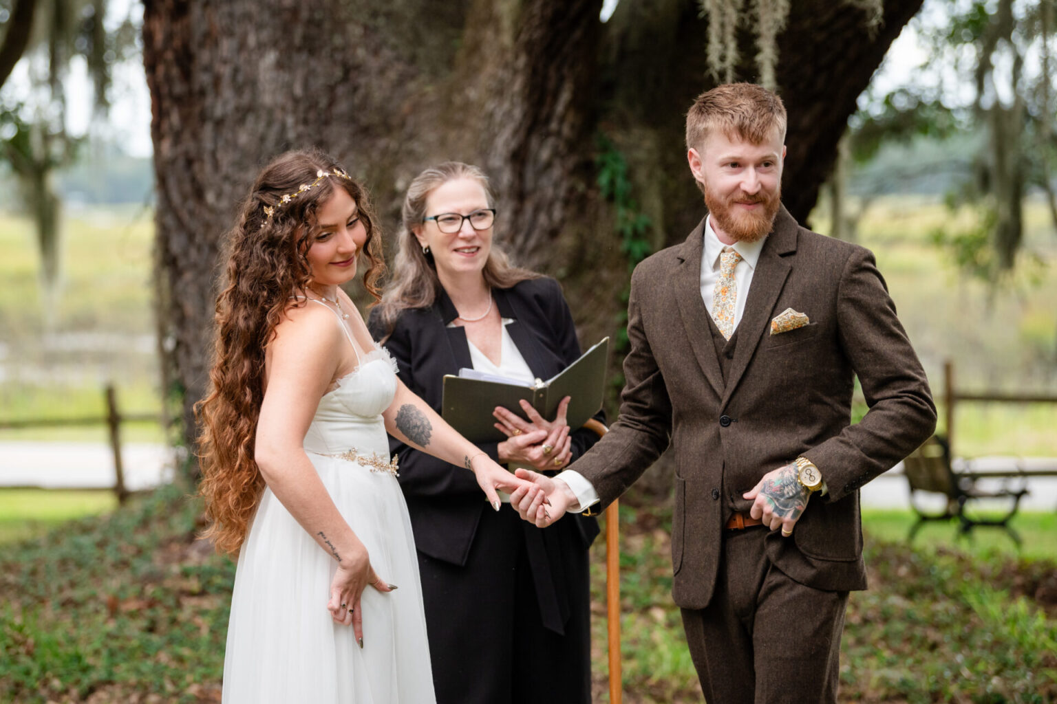 Elopement Wedding A bride and groom joyfully hold hands during an outdoor wedding ceremony. The bride wears a white dress and floral headpiece, and the groom dons a brown suit with a gold tie. An officiant with glasses stands behind them, holding a book. They are in front of a large tree. Elopements Inc