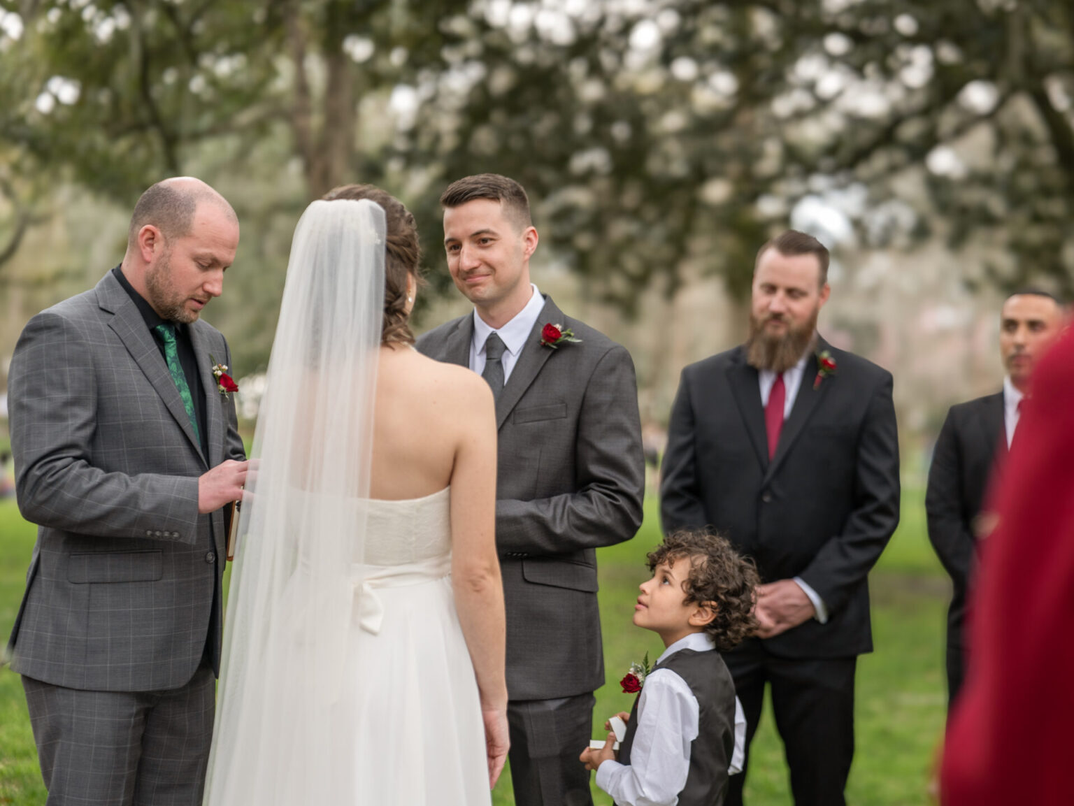 Elopement Wedding A bride and groom stand facing each other during an outdoor wedding ceremony. The groom wears a gray suit and smiles at the bride, who is in a white strapless dress with a veil. A bald officiant in a gray suit reads from a book. A young boy in a vest holds a red rose, looking up at the groom. Elopements Inc