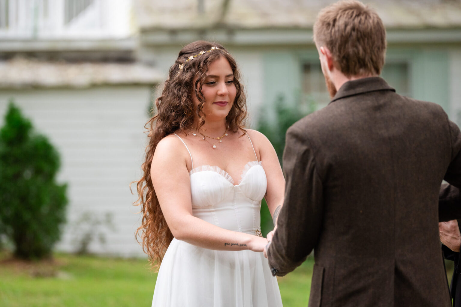 Elopement Wedding A woman in a white wedding dress with curly hair holds hands with a man in a brown suit. They stand outside on grass, in front of a white building. The woman wears a floral hairpiece and delicate jewelry, looking at the man with a soft expression. The scene appears serene and intimate. Elopements Inc