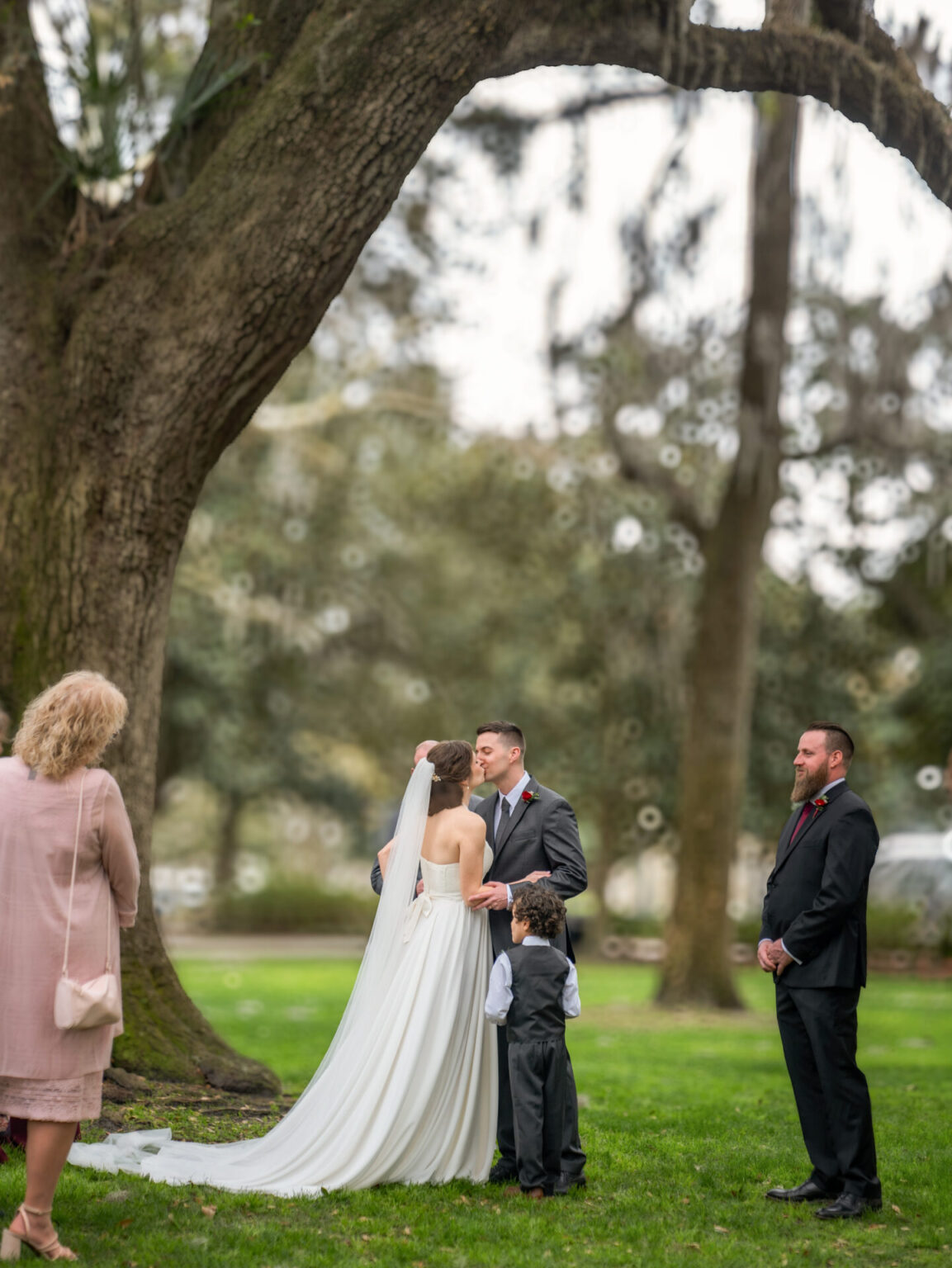 Elopement Wedding A bride and groom kiss under a large tree during their outdoor wedding ceremony. The bride wears a strapless white gown with a long veil. A small child stands beside them, looking up. A woman in a pink outfit watches, and a man in a suit stands nearby under the tree canopy. Elopements Inc