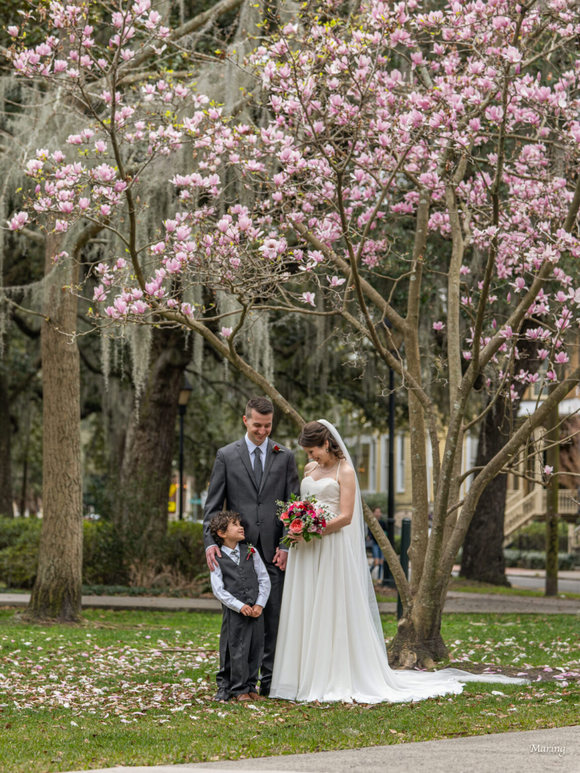 Elopement Wedding A couple in wedding attire stands with a young boy under blooming pink magnolia trees in a park. The groom wears a gray suit, the bride a white gown, and the boy a gray vest with a bow tie. They are looking at each other and smiling. Pink petals are scattered on the grass. Elopements Inc