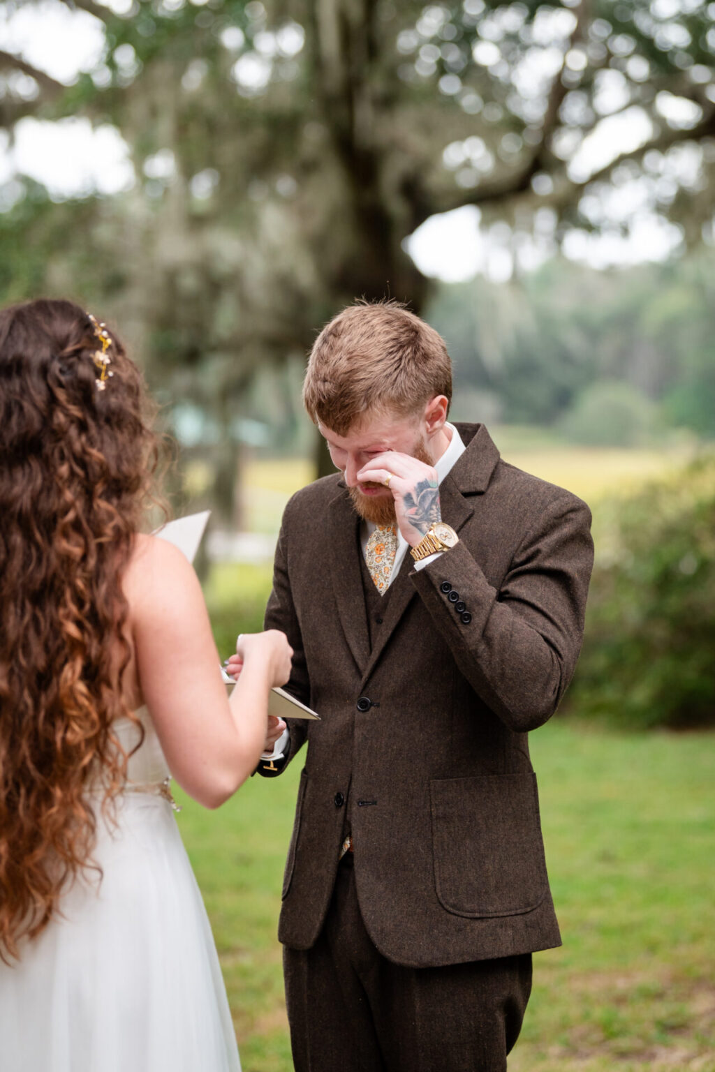 Elopement Wedding A man in a brown suit wipes his eye while a woman in a white dress, with long curly hair, reads from a card. They stand outdoors on grass with a tree in the background. The scene suggests an emotional moment, possibly during a wedding ceremony. Elopements Inc