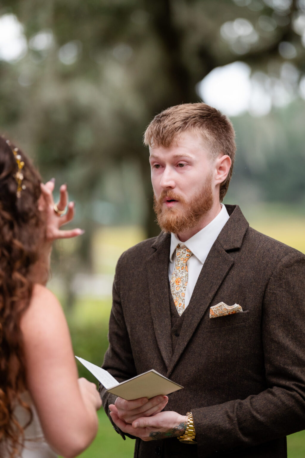 Elopement Wedding A person with a beard and brown hair stands outdoors, wearing a brown suit, floral tie, and pocket square. They hold an open book, appearing engaged. Another person with long hair is partially visible, facing them. The background shows blurred greenery. Elopements Inc