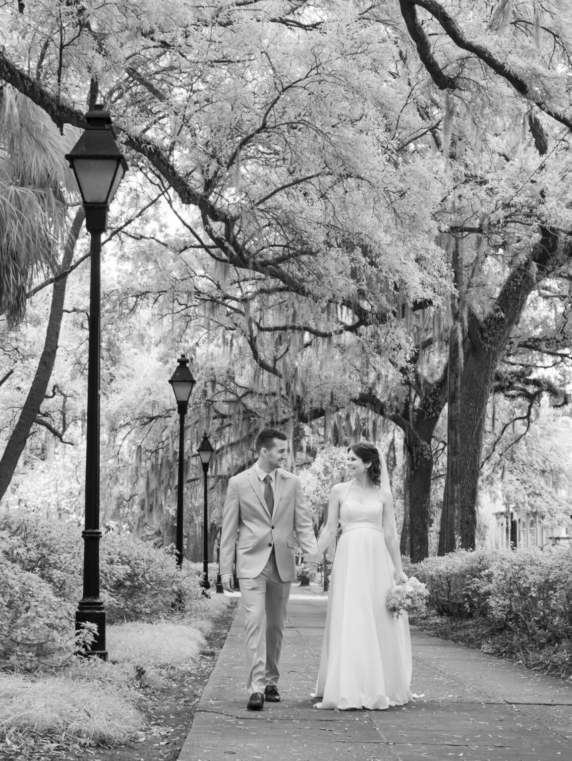 Elopement Wedding A couple in wedding attire walks hand in hand along a path lined with street lamps and large trees draped with Spanish moss. The bride wears a long white dress and holds a bouquet, while the groom is in a light-colored suit. The setting is serene and romantic, captured in black and white. Elopements Inc