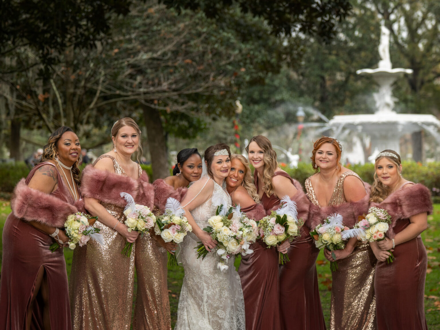 Elopement Wedding A bride in a white dress stands with six bridesmaids in elegant, burgundy and gold gowns. They hold bouquets of white and pink flowers. The group poses outdoors with a large, ornate fountain visible in the background, amidst lush greenery and trees. Elopements Inc