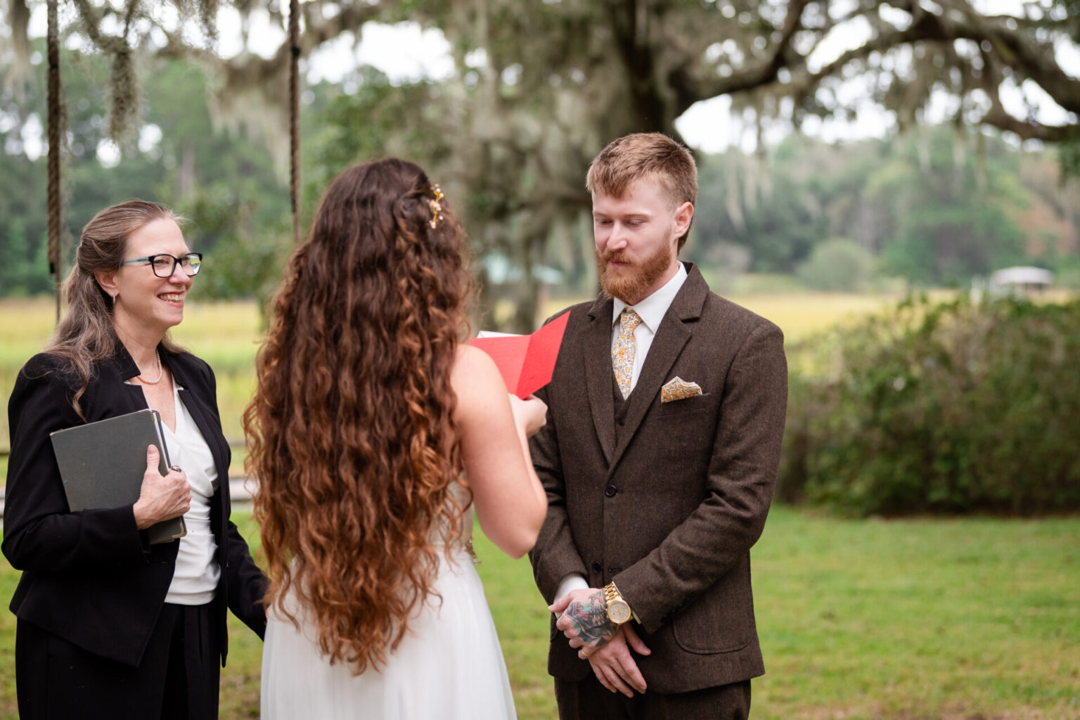 Elopement Wedding A bride in a white dress with long curly hair reads vows from a red booklet to a groom in a brown suit with a floral tie. A woman officiant in glasses and a black suit holds a black folder. They stand outdoors with mossy trees and greenery in the background. Elopements Inc